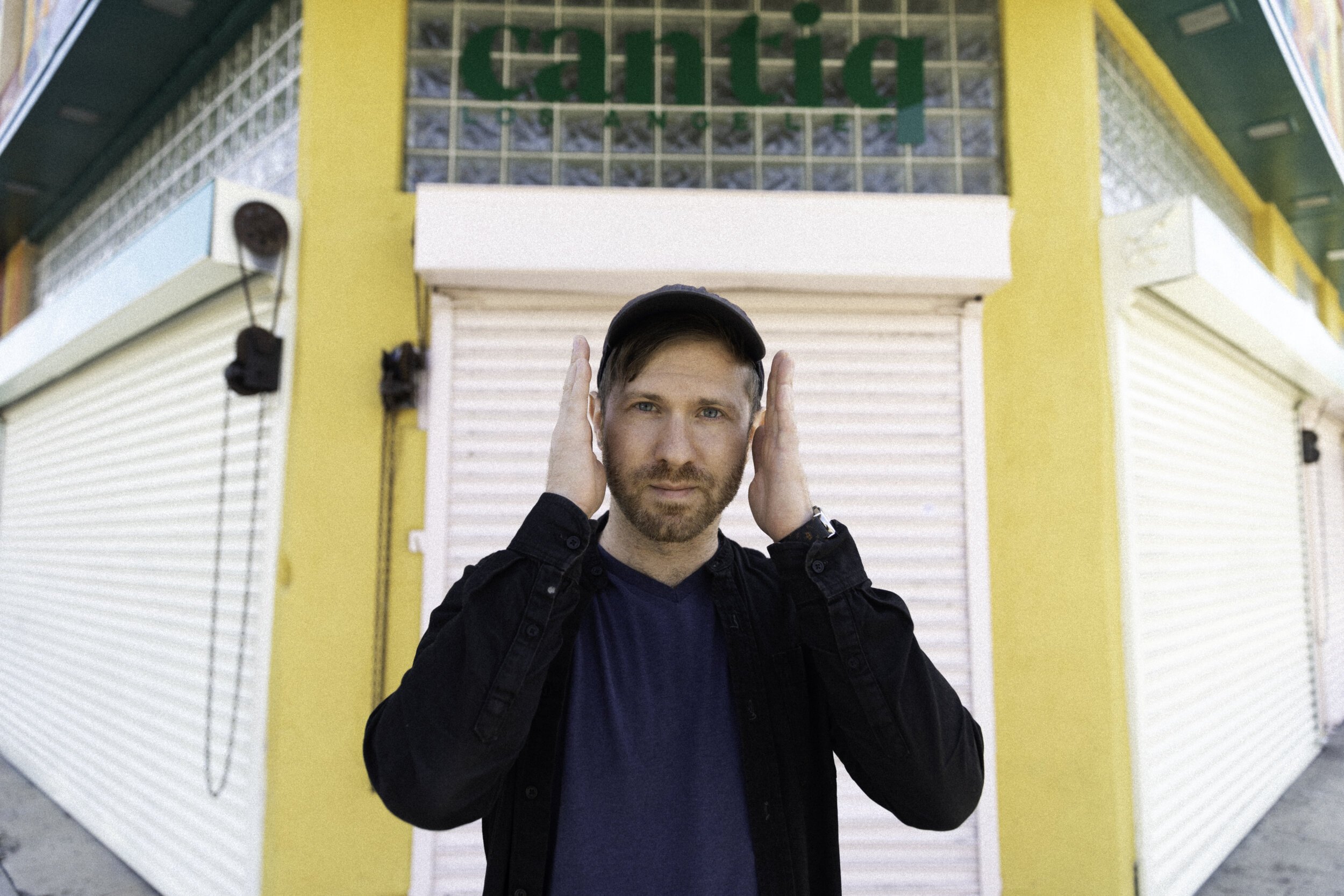 A man with a beard and cap standing in front of a closed storefront with a yellow and white exterior while holding his hands near his ears.