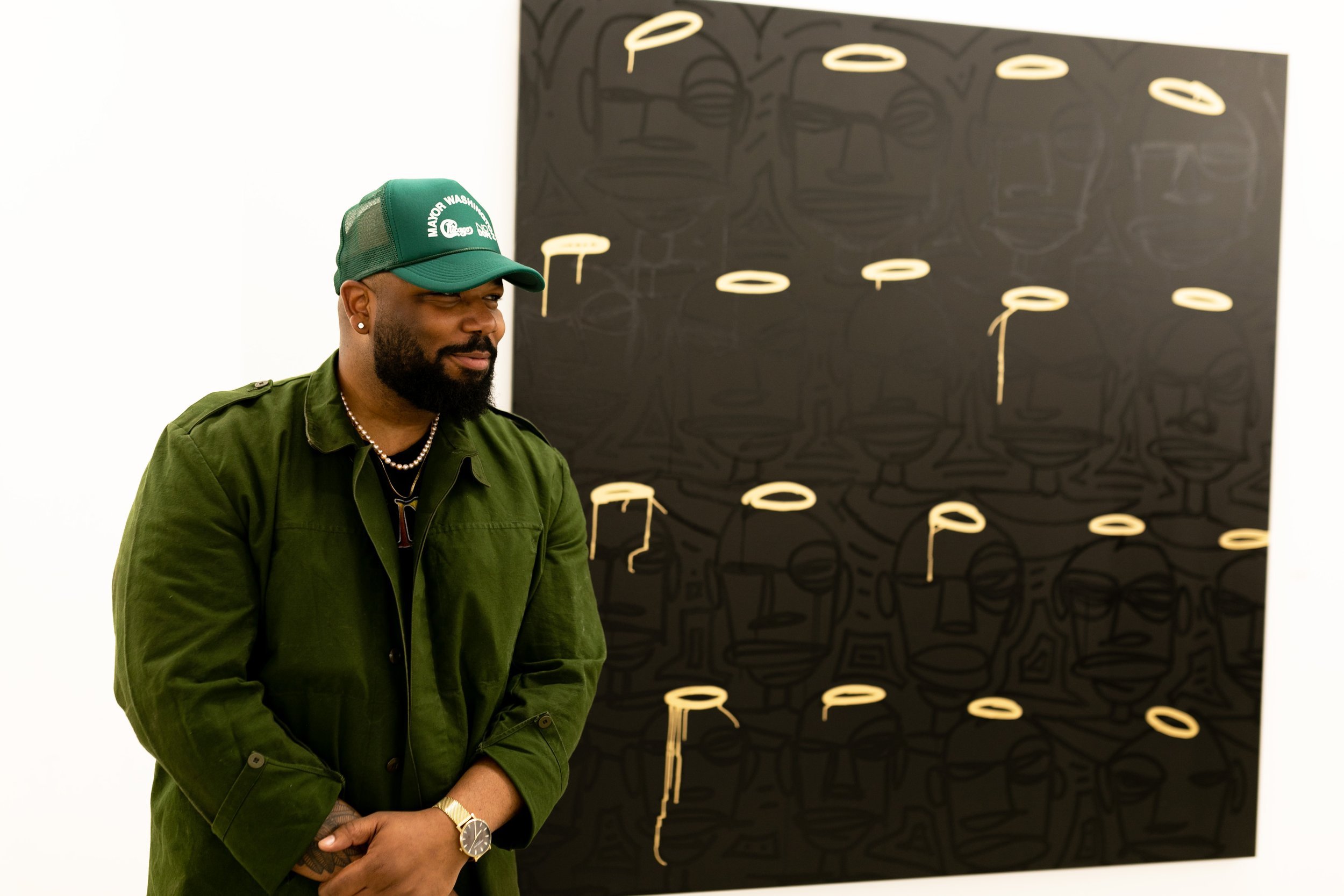 Brian Dovie Golden with a beard and earrings wearing a green jacket and green cap standing in front of black artwork with faint line drawings of faces and glasses