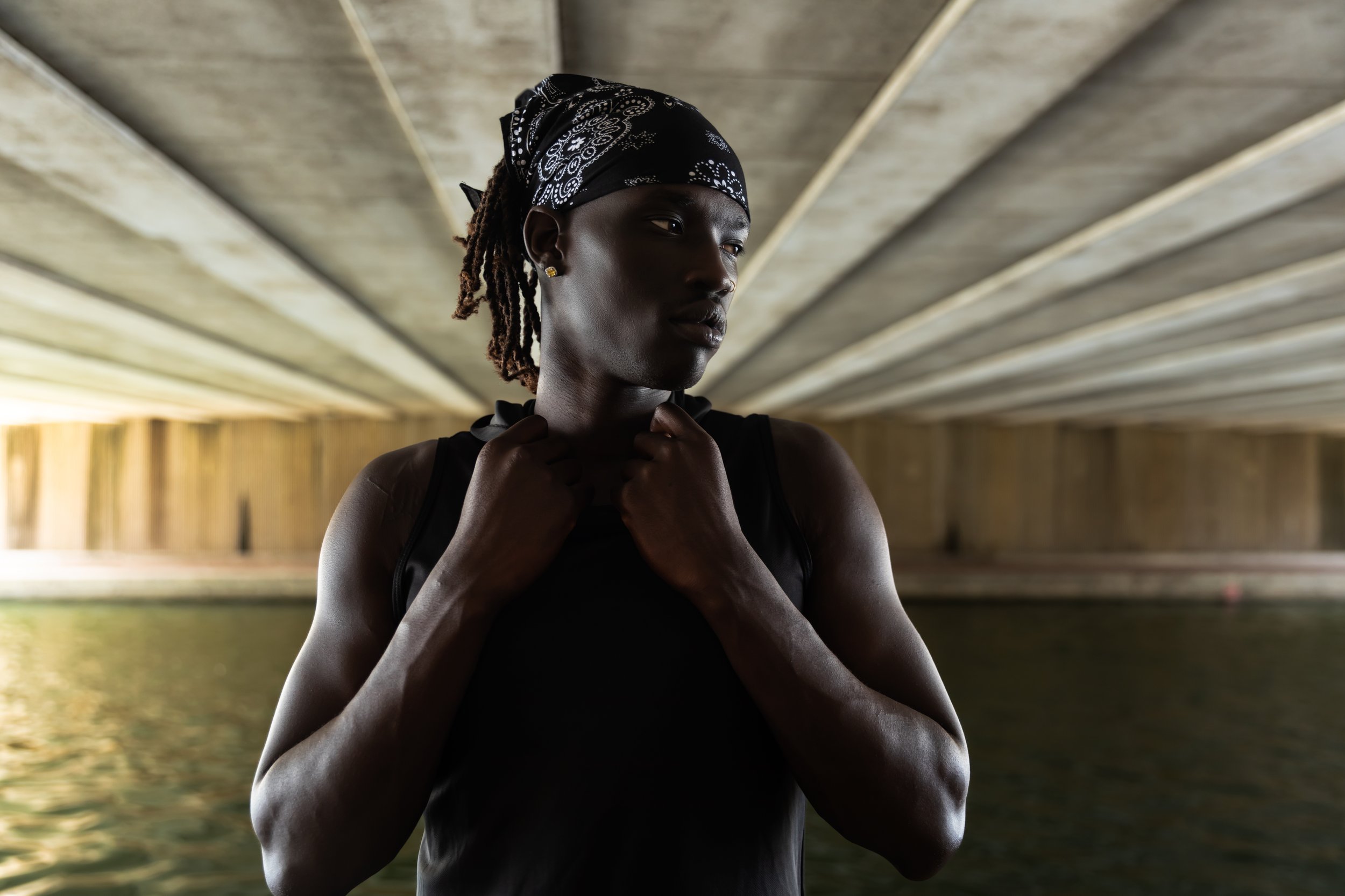 A woman with dark skin and dreadlocks wearing a black bandana and a black sleeveless top, standing under a concrete bridge with water below.