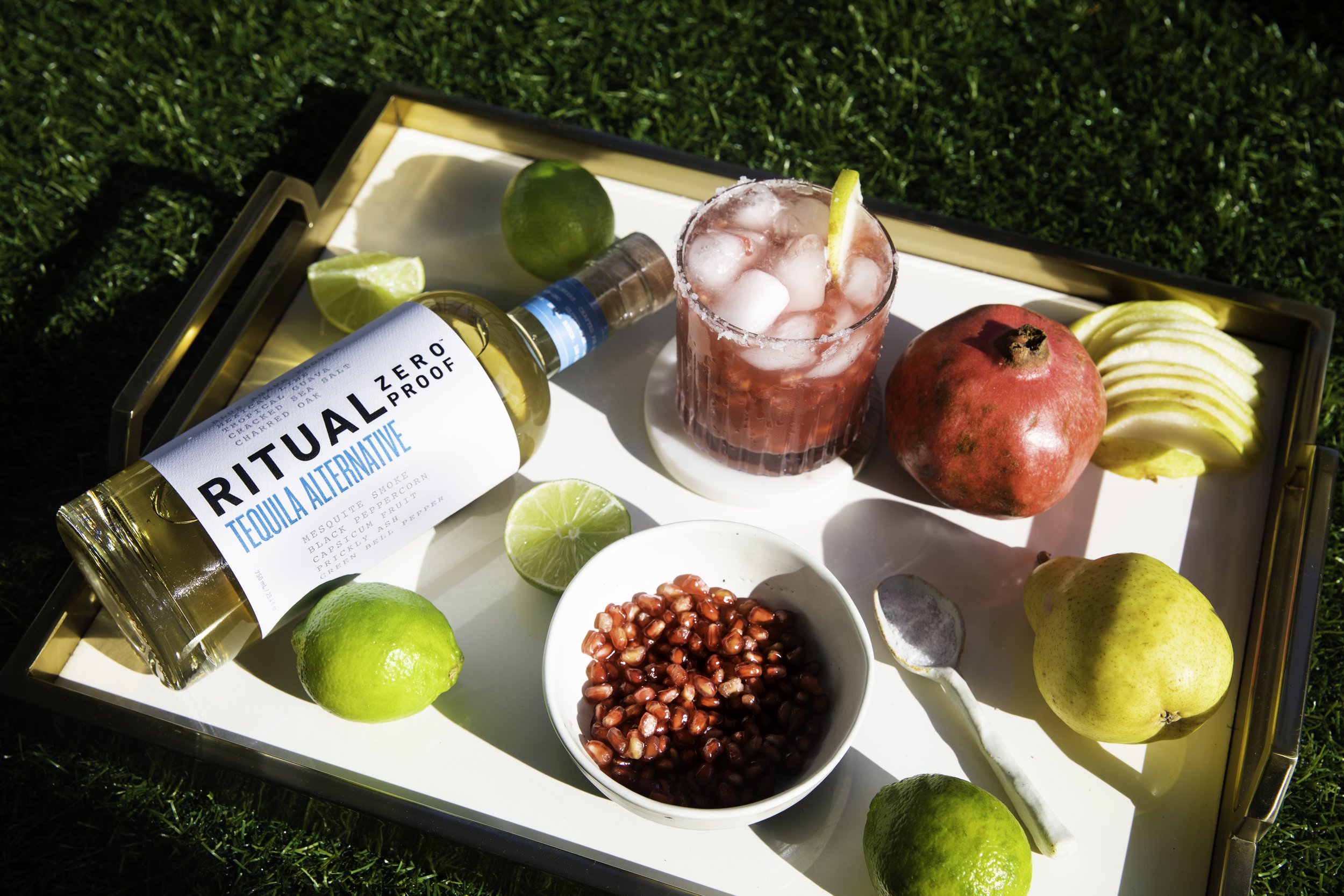 A tray containing a bottle of Ritual Zero Proof tequila alternative, a pomegranate, a glass of pink cocktail with ice and lemon slice, several lime and lemon wedges, a bowl of pomegranate seeds, and sliced green apples on a grassy surface.