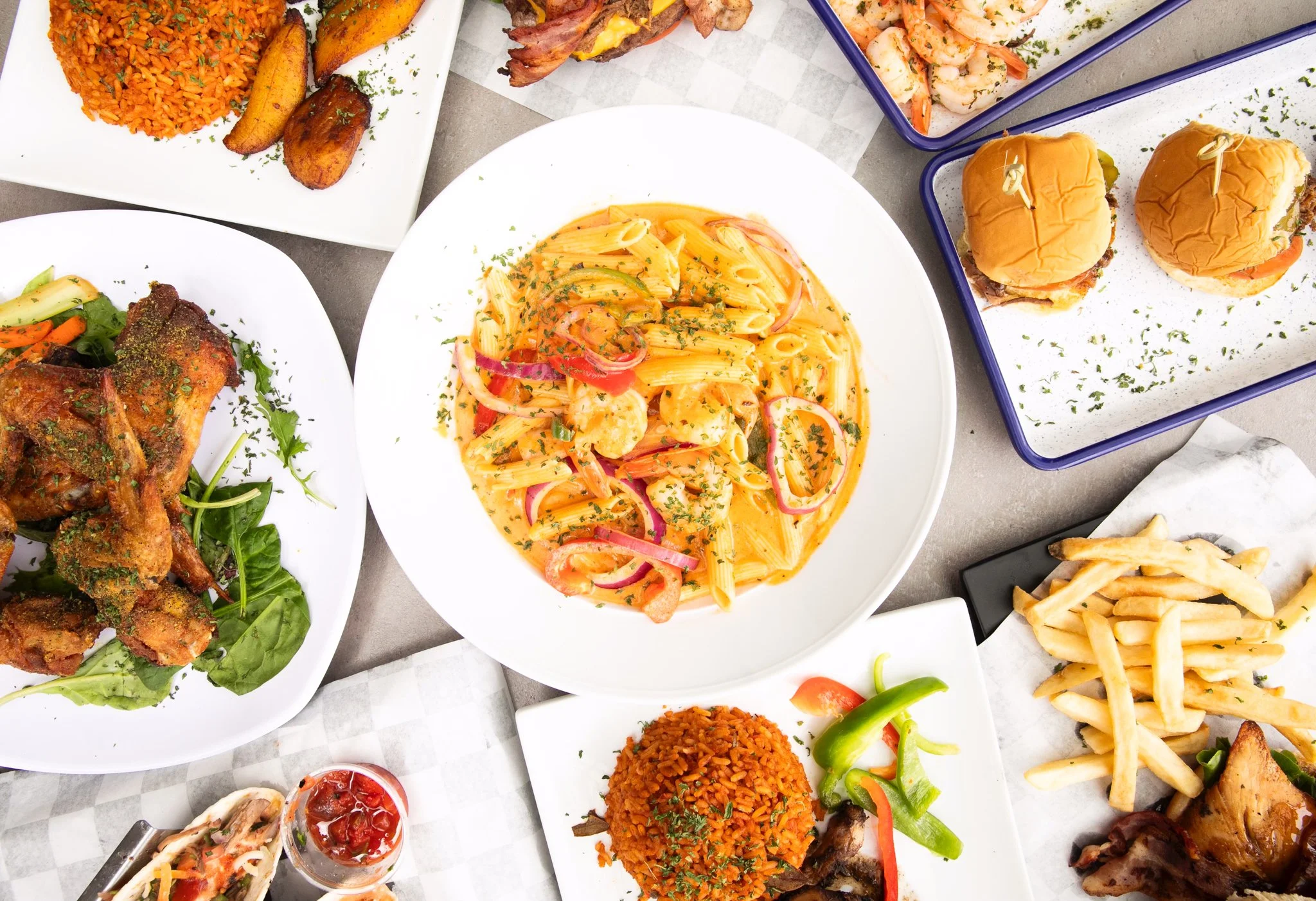 A variety of dishes on a table, including pasta with shrimp and vegetables, fried chicken, rice, fries, burger sliders, shrimp, and a small salad with pepper sauce.