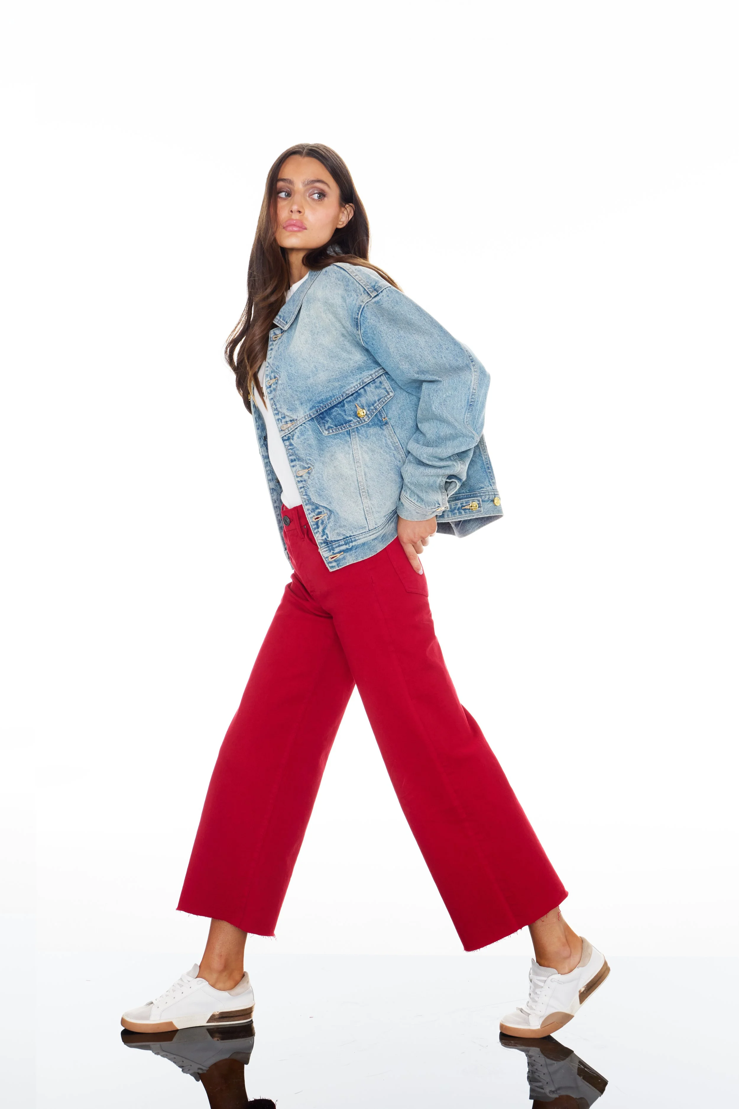 Young woman in casual outfit with denim jacket, white shirt, red wide-leg pants, and white sneakers, posing with one hand on her hip against a white background.