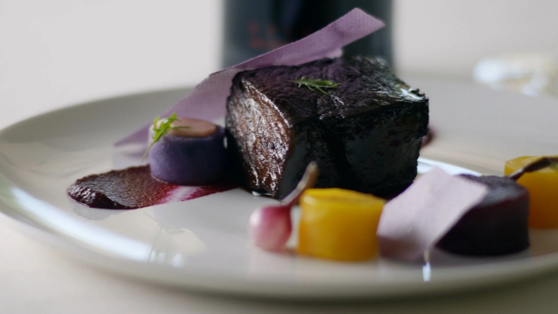 A plated gourmet dish featuring a piece of seared meat with a glossy dark glaze, garnished with a small green herb and accompanied by purple and yellow vegetable elements, presented artistically with sauces and decorative paper.