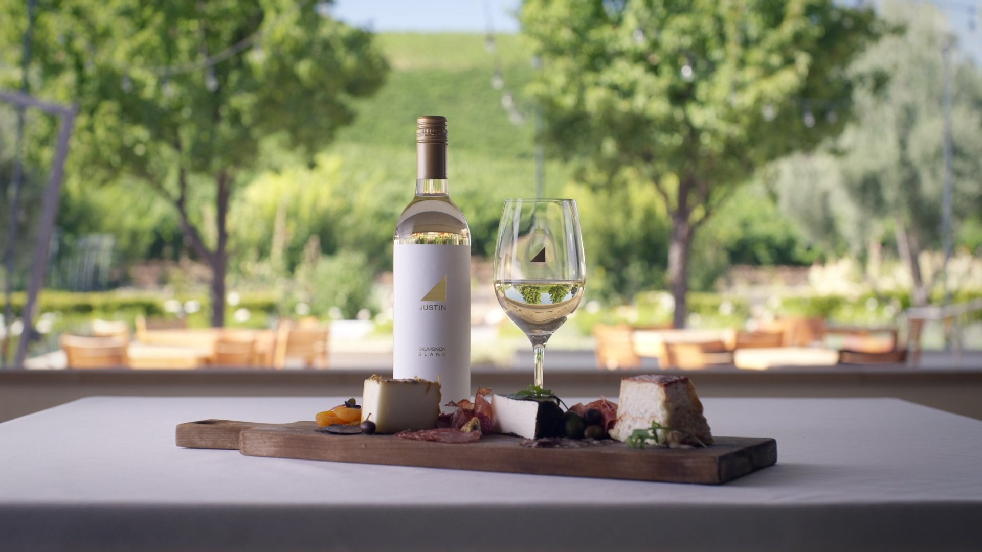 A wine bottle labeled 'Justin' and a filled wine glass sit on a wooden serving board with assorted cheese, cured meats, and grapes, set on a white tablecloth with outdoor greenery in the background.