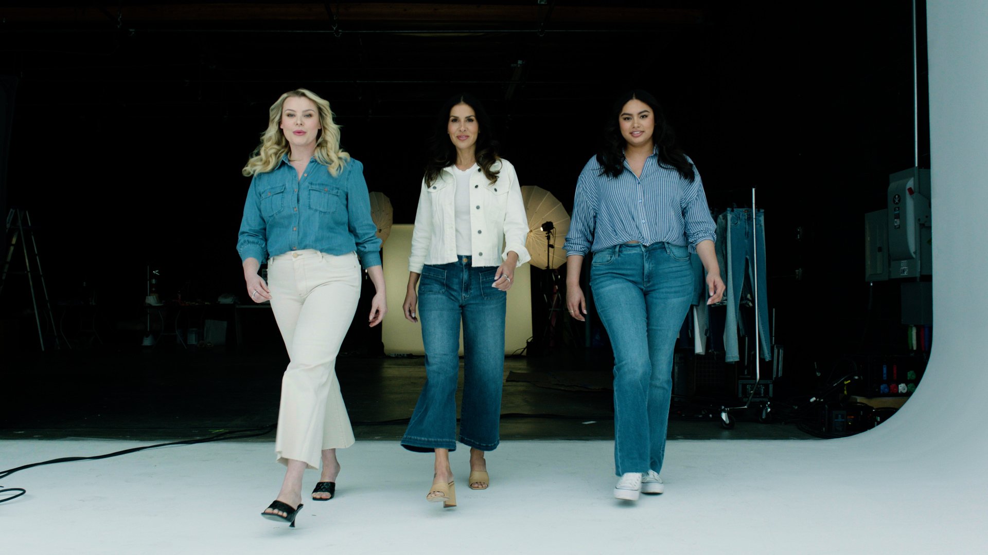 Three women walking towards the camera in a photo studio with studio lights and equipment in the background. They are wearing casual denim outfits.
