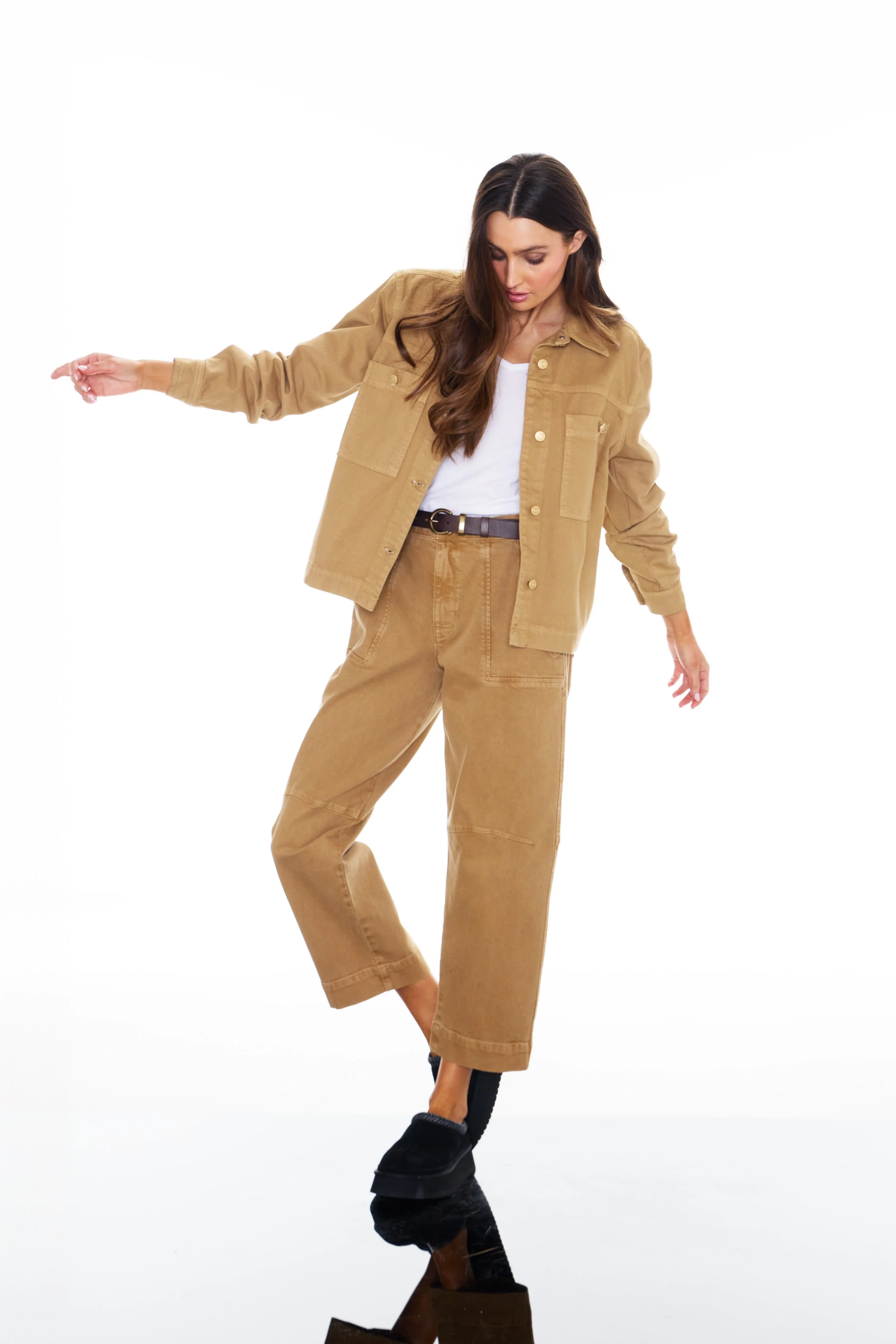 A woman dressed in a light brown jacket and pants, wearing black shoes, balancing on one foot against a white background.