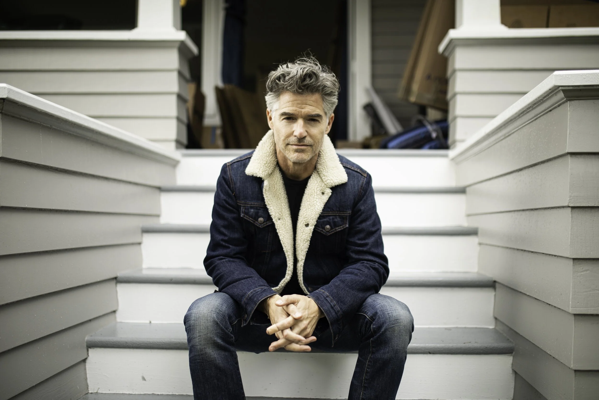 A man with gray hair, wearing a dark jacket with a shearling collar, sits on the steps of a porch with a serious expression, in front of a house with visible clutter inside.