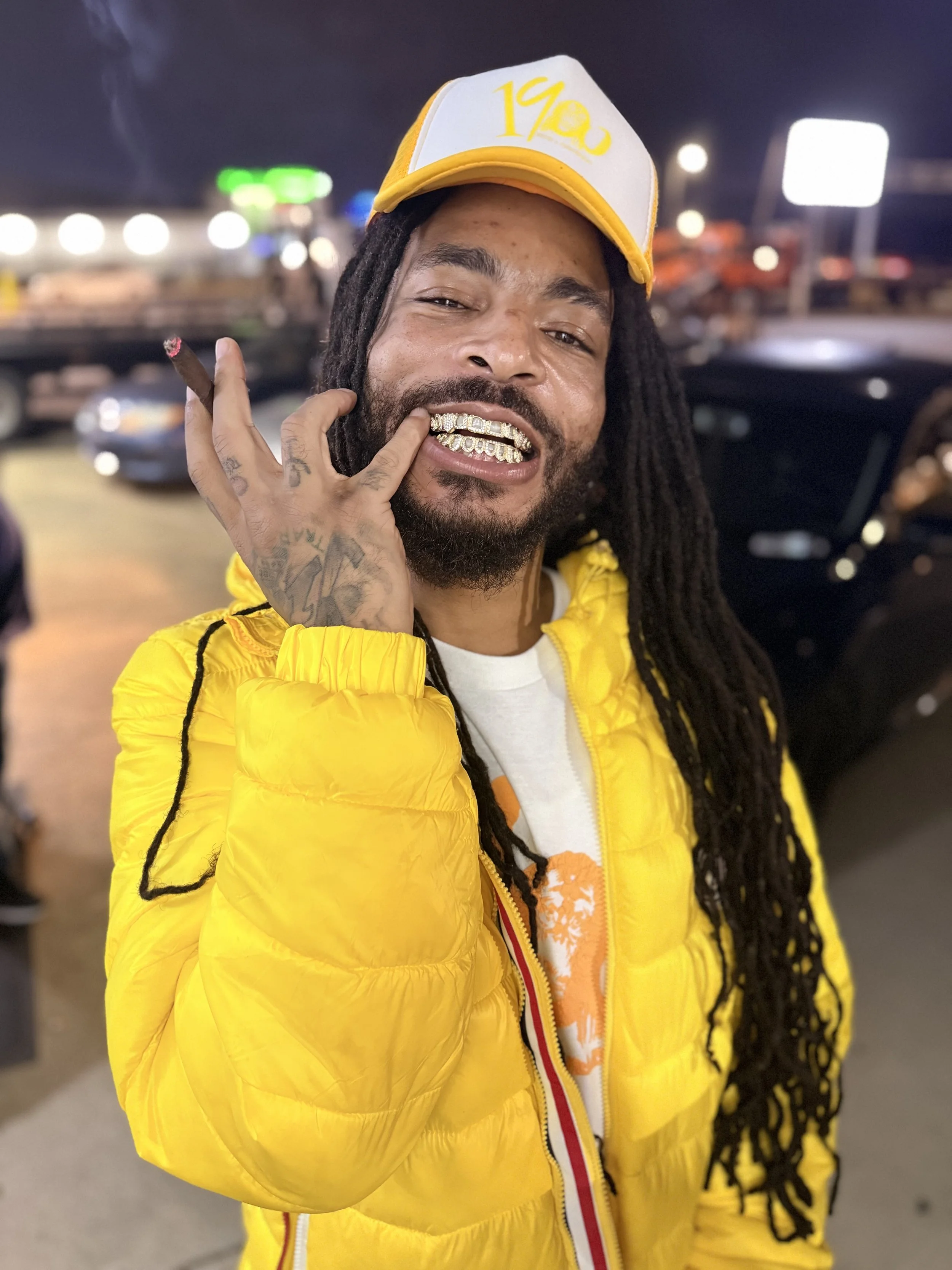 Man with long dreadlocks, wearing a yellow puffer jacket and a white cap with yellow lettering, showing gold teeth and holding a lit cigar in his mouth, at night with blurred lights and cars in the background.
