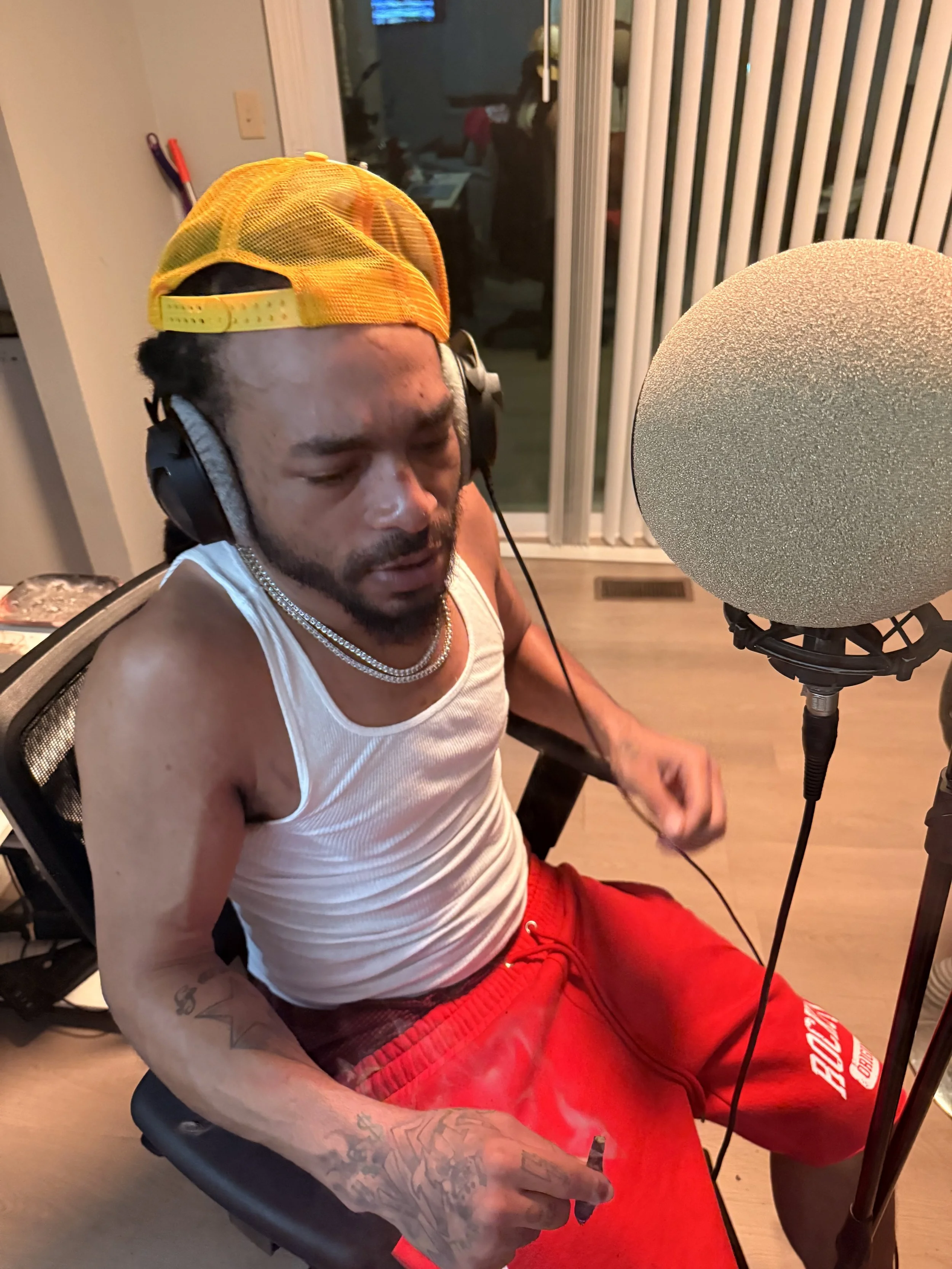 A man wearing a yellow trucker hat, white tank top, red shorts, and headphones is sitting in front of a microphone in a recording studio.
