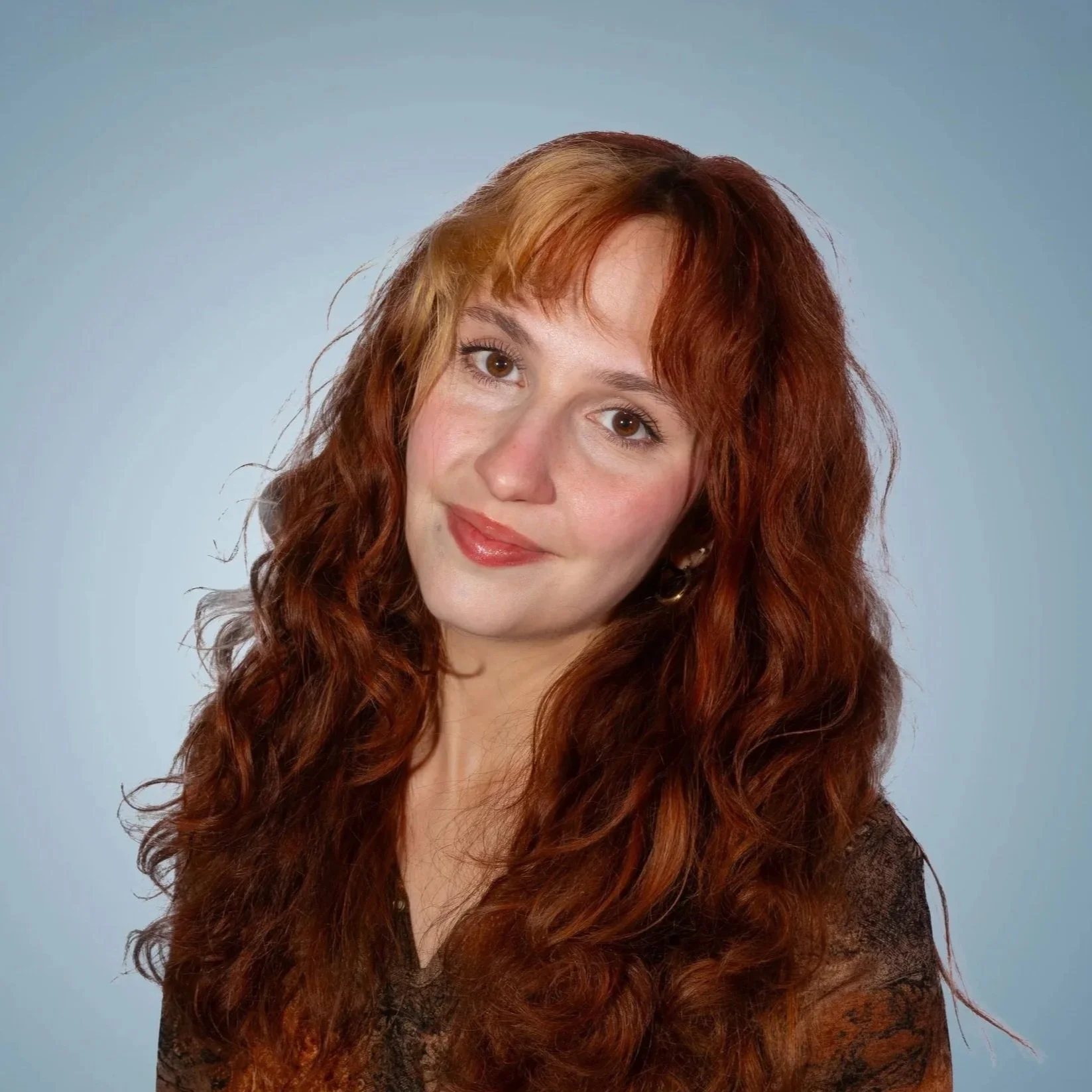 Lamija Memidzan, Salt Lake City-based creative photographer with long, curly red hair and brown eyes, wearing a dark top, smiling softly against a light blue background.