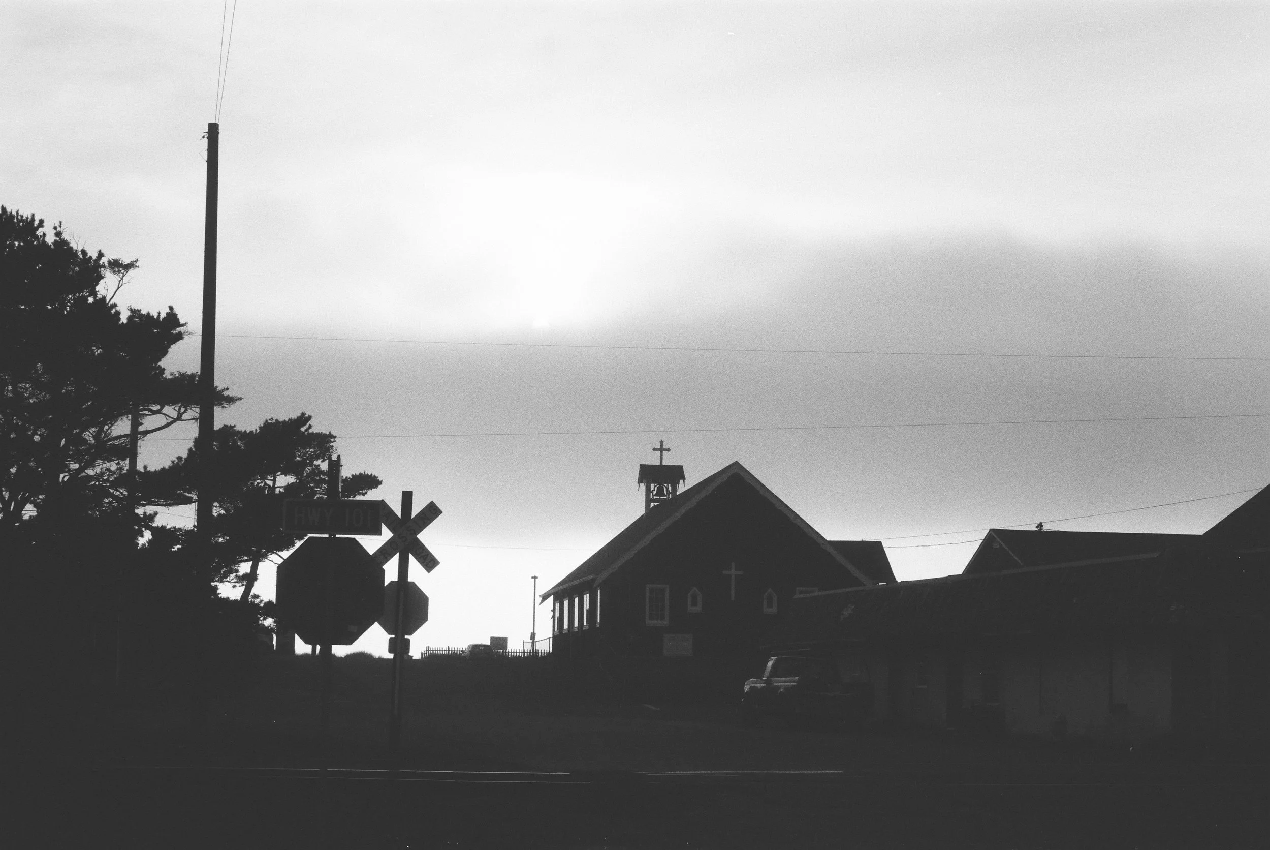 Black and white photo of a church with a steeple topped by a cross, a church bell, and houses, with a tree on the left and power lines overhead, under a cloudy sky.