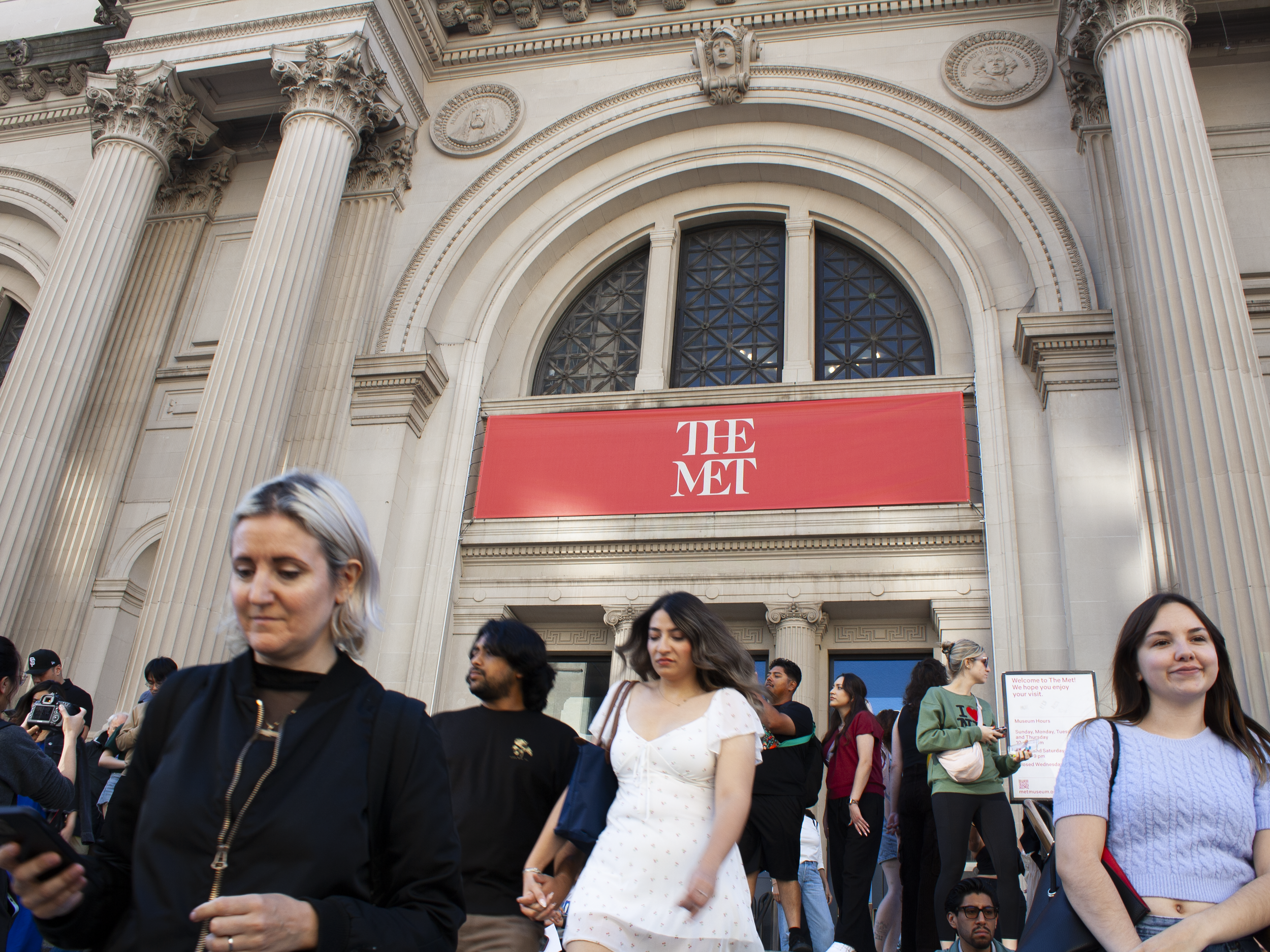 Crowd of people walking outside of the Metropolitan Museum of Art, with the museum's ornate architectural facade and a red sign reading 'THE MET' visible in the background.