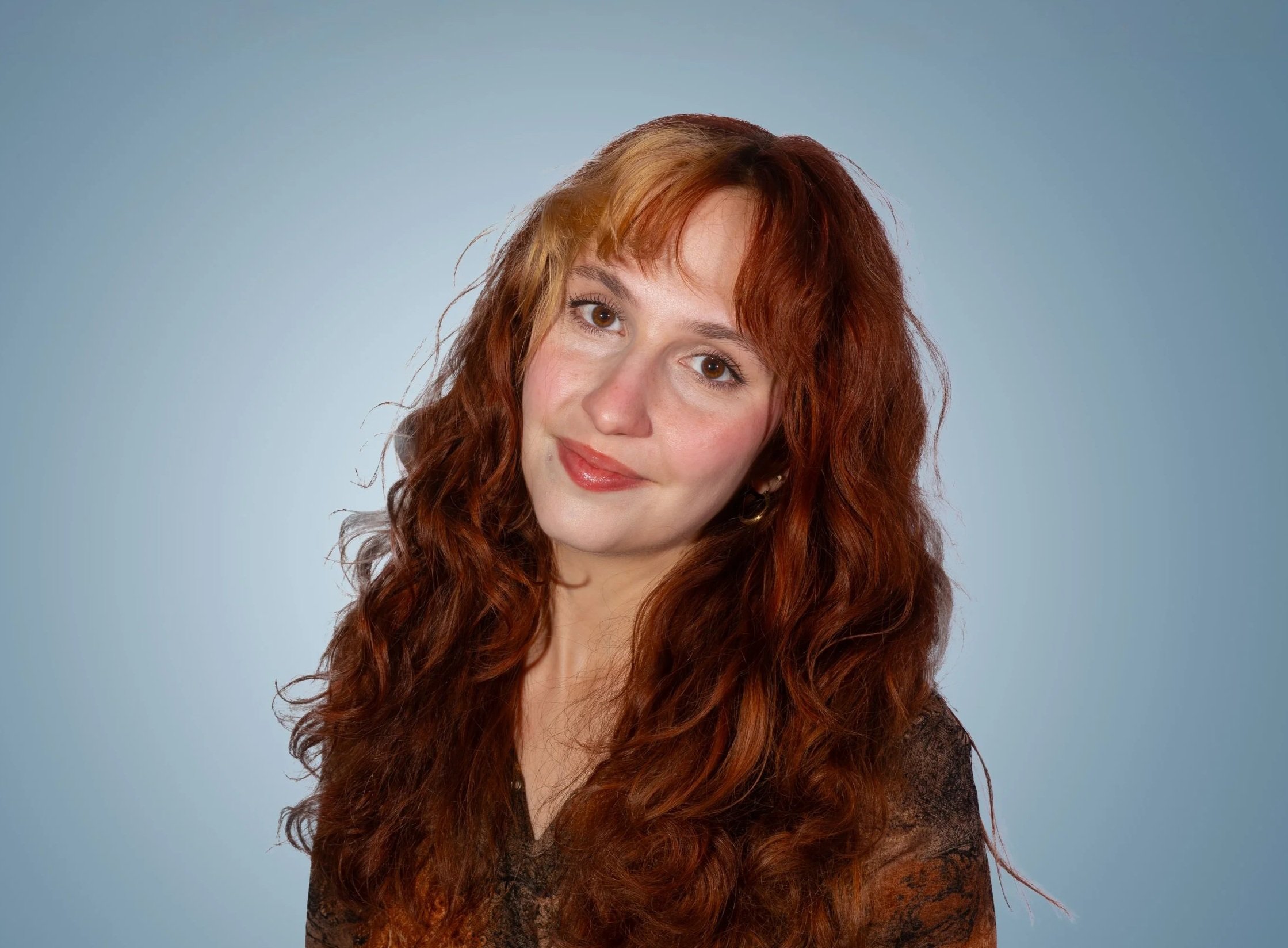 Lamija Memidzan, Salt Lake City-based creative photographer with long, curly red hair and brown eyes, wearing a dark top, smiling softly against a light blue background.