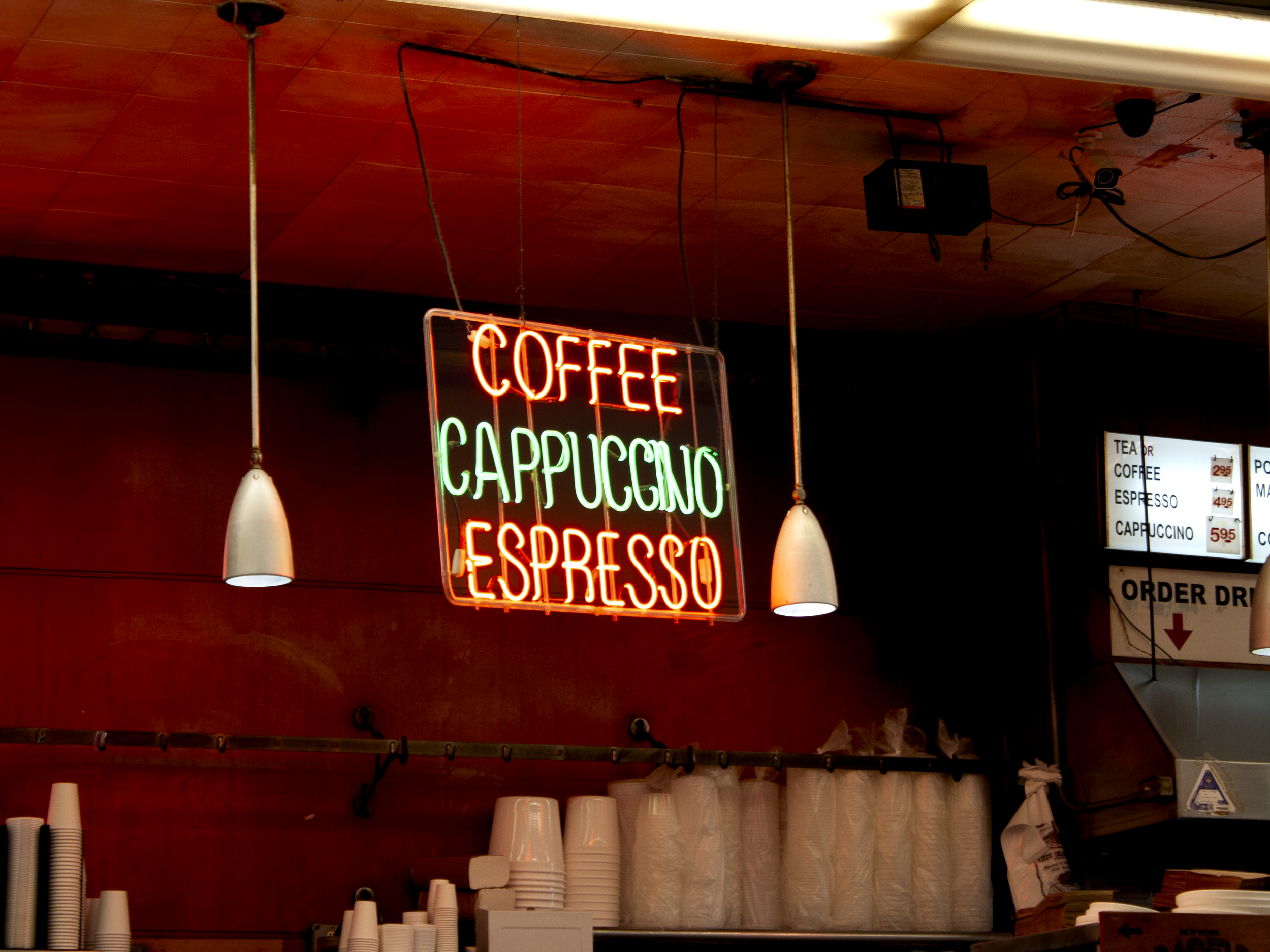 Neon sign displaying the words 'COFFEE,' 'CAPPUCCINO,' and 'ESPRESSO' hanging from the ceiling of a coffee shop, with cups and coffee-related items below.