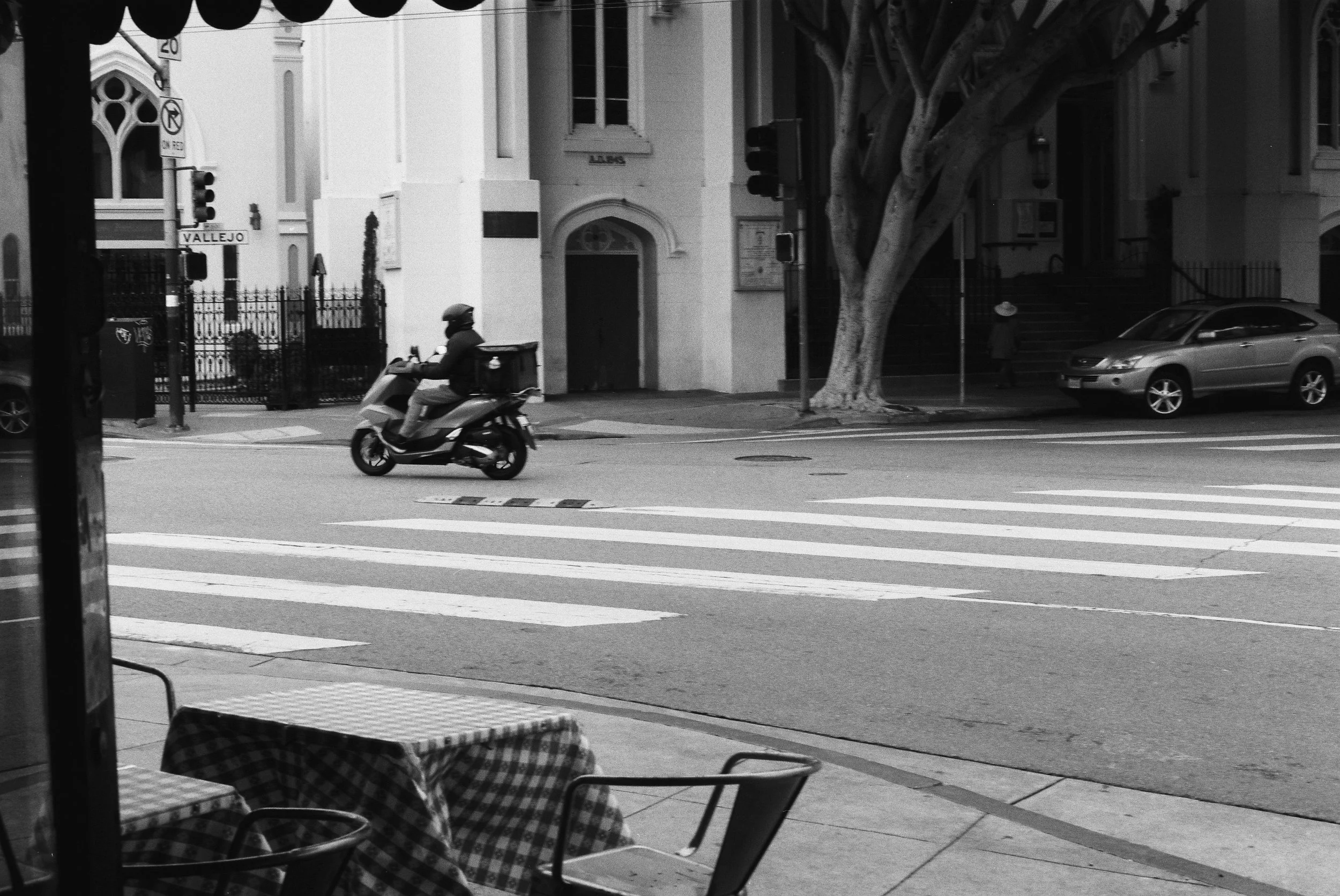 Black and white photo of a city street viewed from inside a cafe. A person riding a scooter is crossing the street. There are tall trees, parked cars, and buildings in the background, with street signs and traffic lights visible.