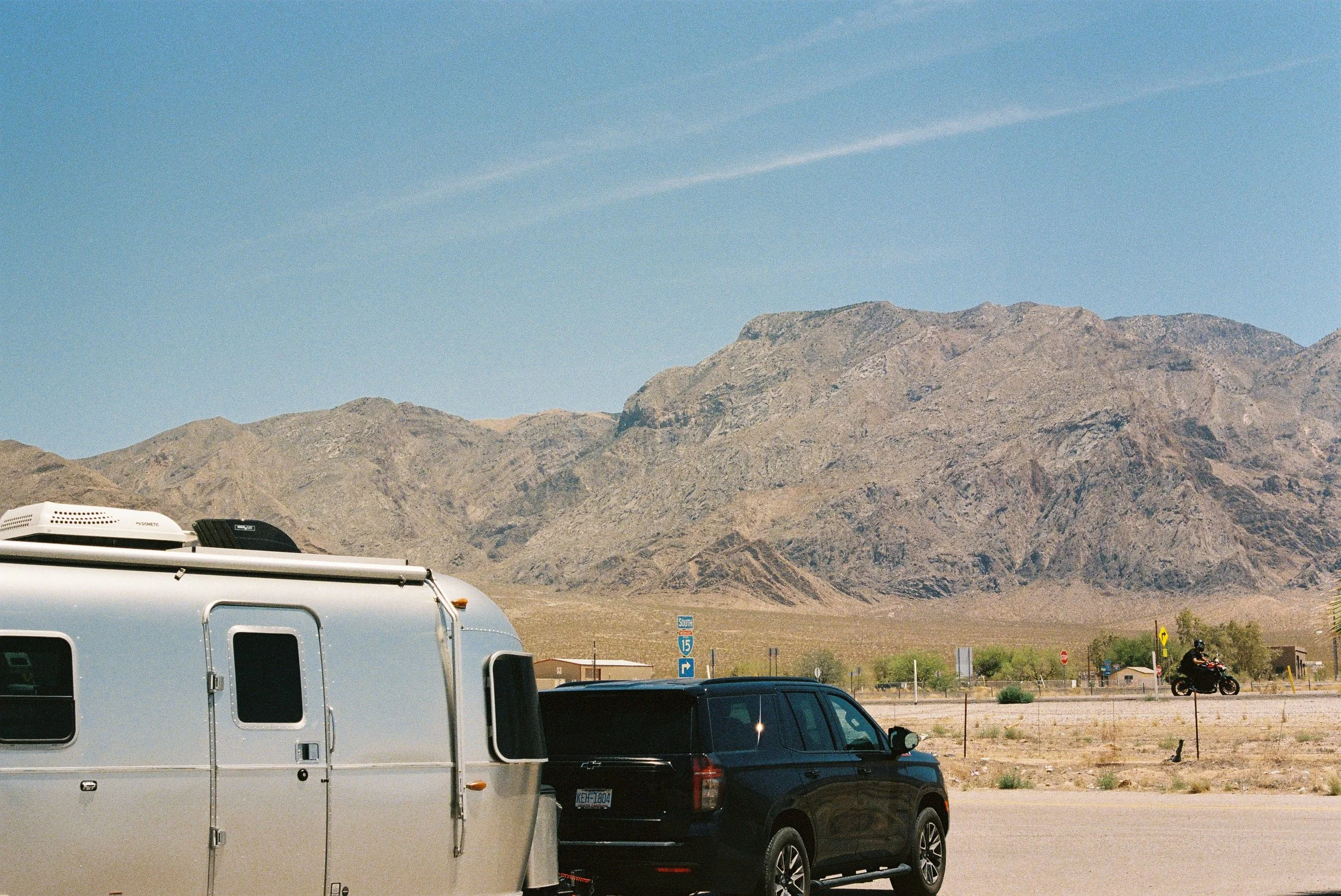 A parking lot with several vehicles, including a van and a pickup truck, with a desert mountain landscape in the background under a blue sky.