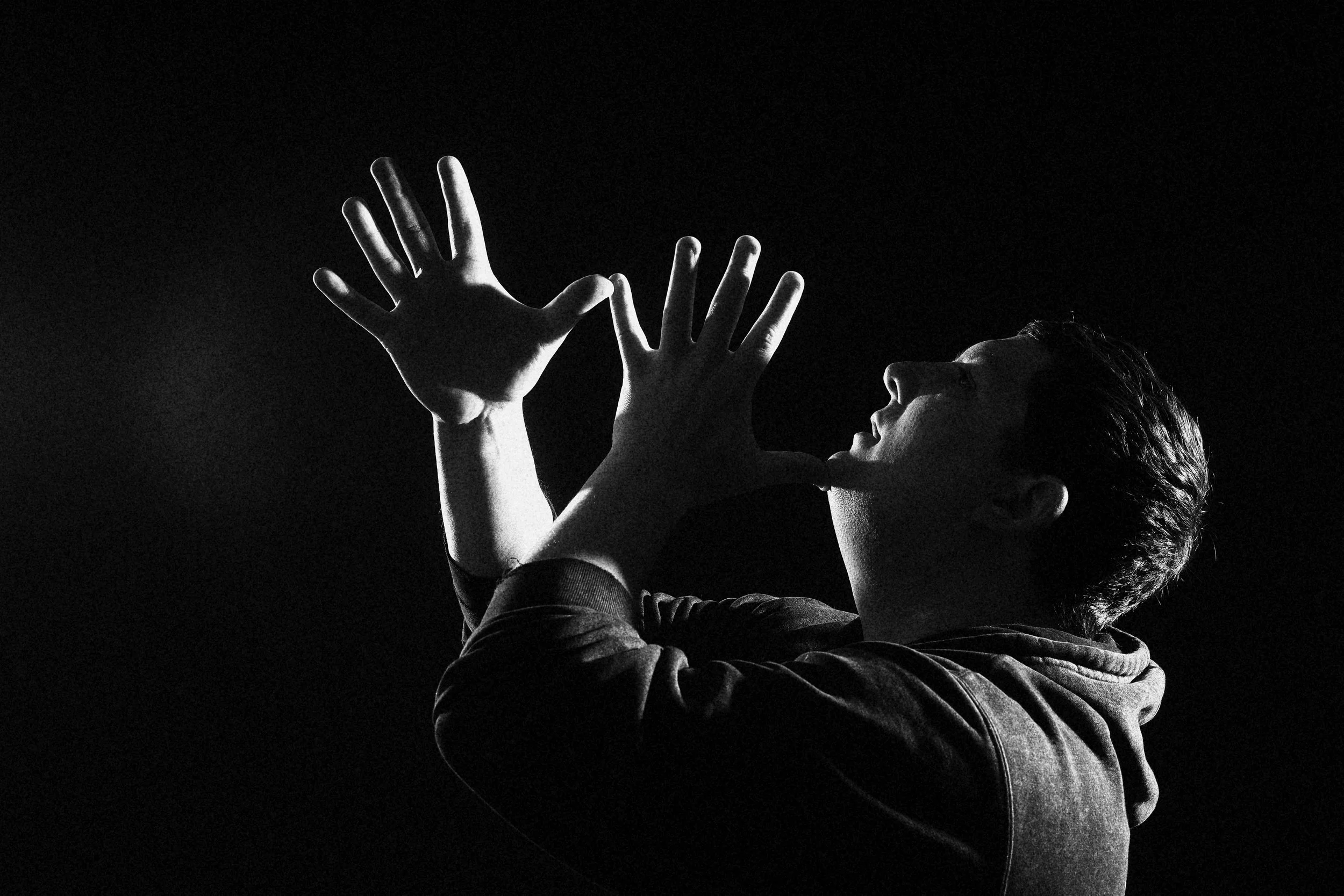 Black and white photo of a person with short hair raising hands against a dark background.