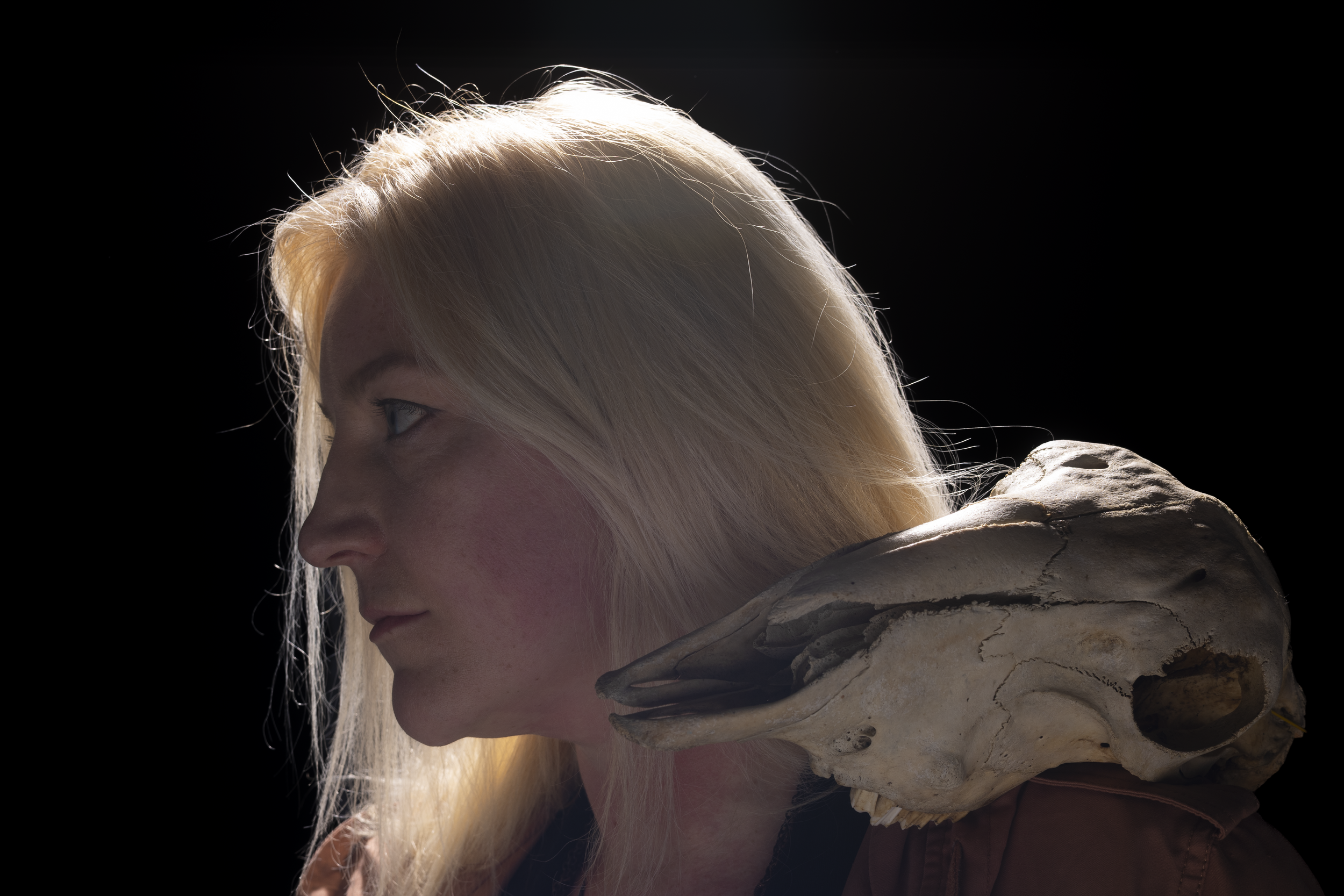 Profile of a blonde woman with light-colored hair, shown in side profile, against a dark background, with a large animal skull with horns resting on her shoulder.