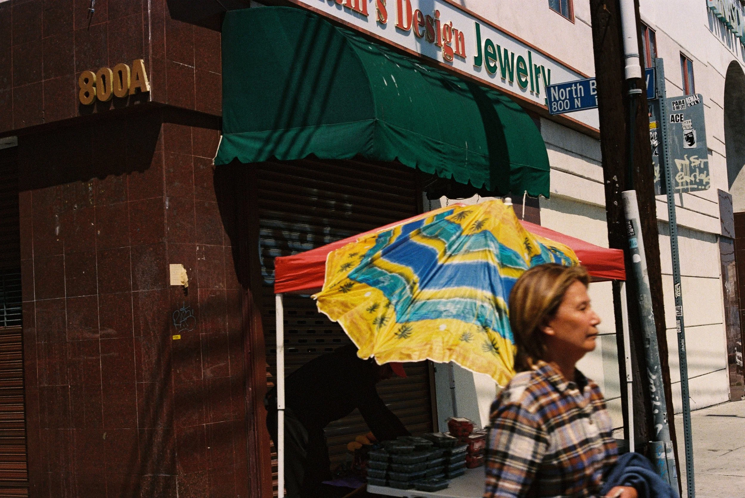 Storefront with green awning, sidewalk vendor display, and a person in a plaid shirt walking by