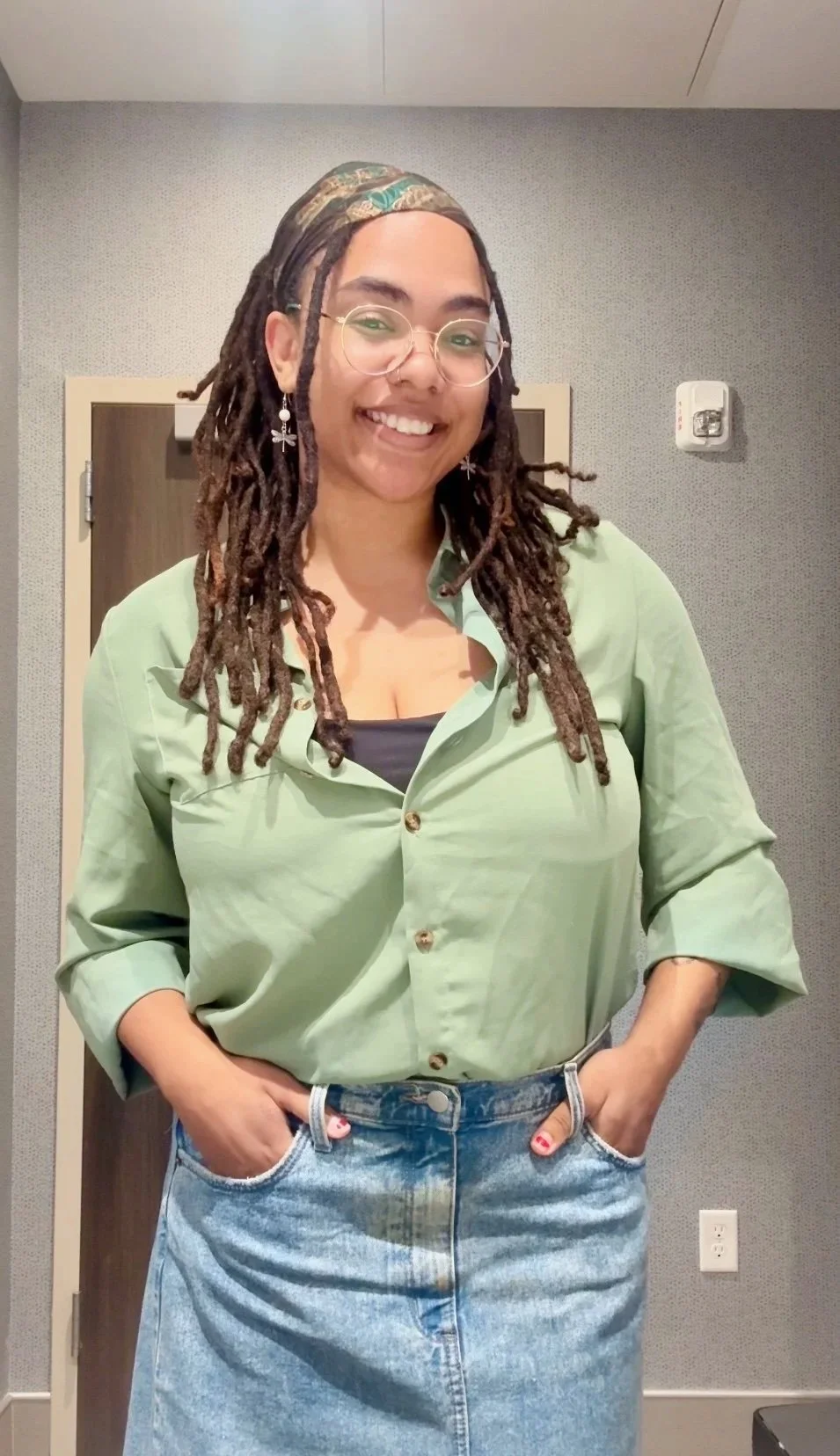 A woman with glasses and earrings smiling, with her hands in her pockets, standing in front of a gray wall and a closed door.