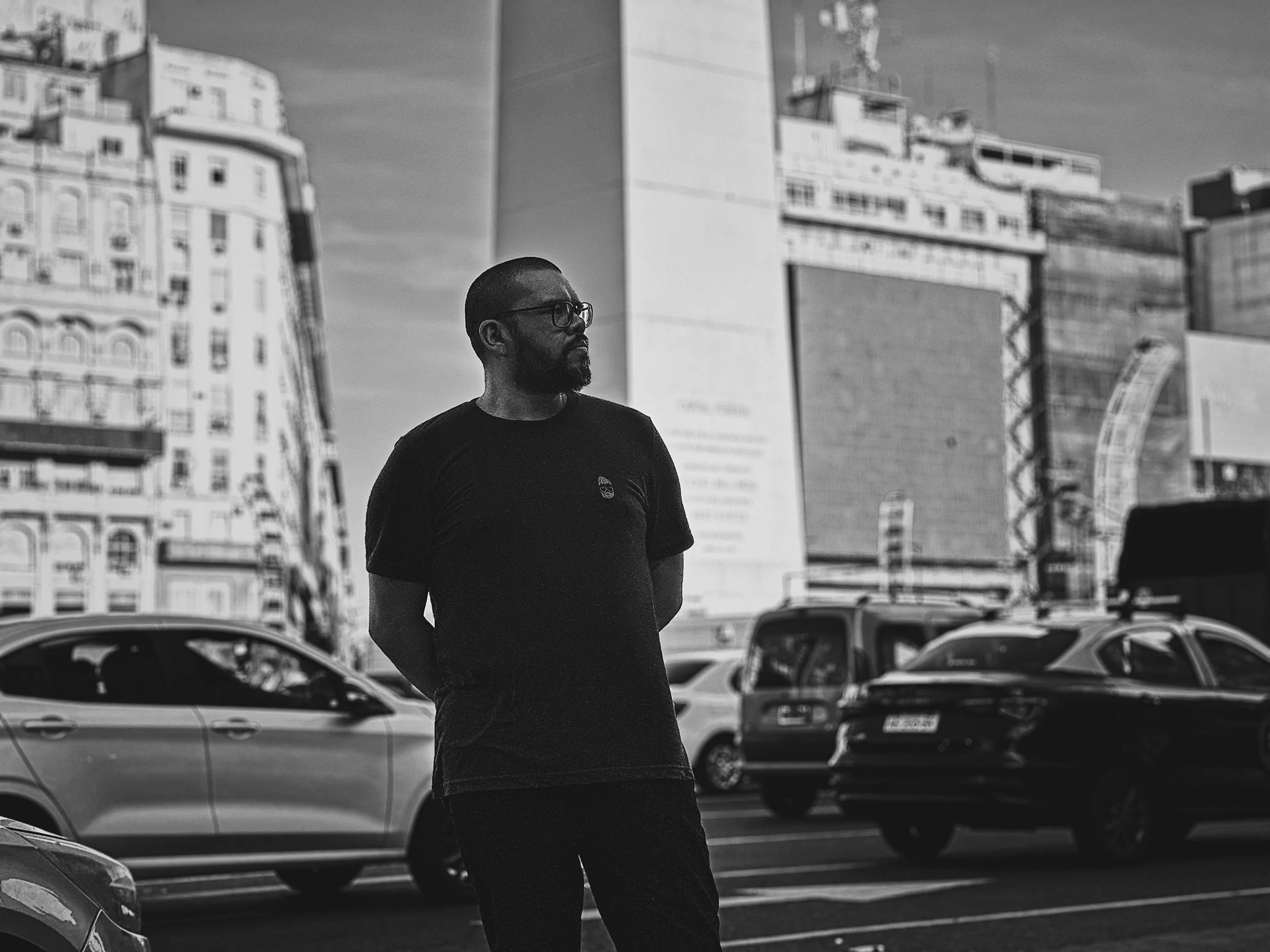 A man with glasses and a beard stands with his hands behind his back on a city street next to parked cars, with tall buildings in the background.