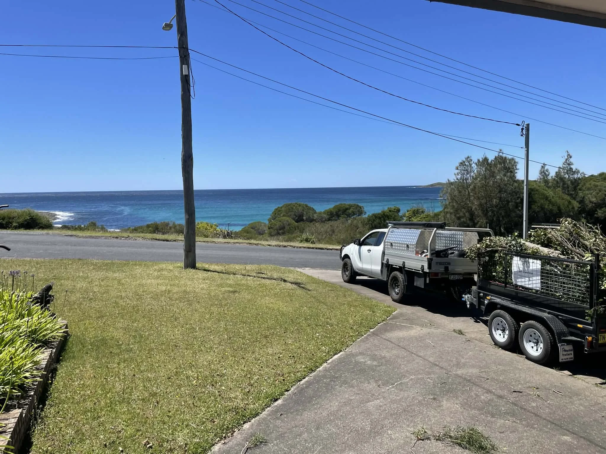 A view of a coastline with clear blue water and a small hill in the distance. In the foreground, there is a truck and a trailer parked on a driveway, with power lines overhead and a grassy area next to the driveway.