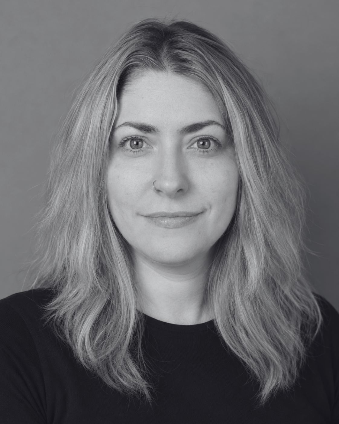 Black and white portrait of a woman with wavy hair, wearing a nose ring and a black shirt, smiling slightly, against a plain background.