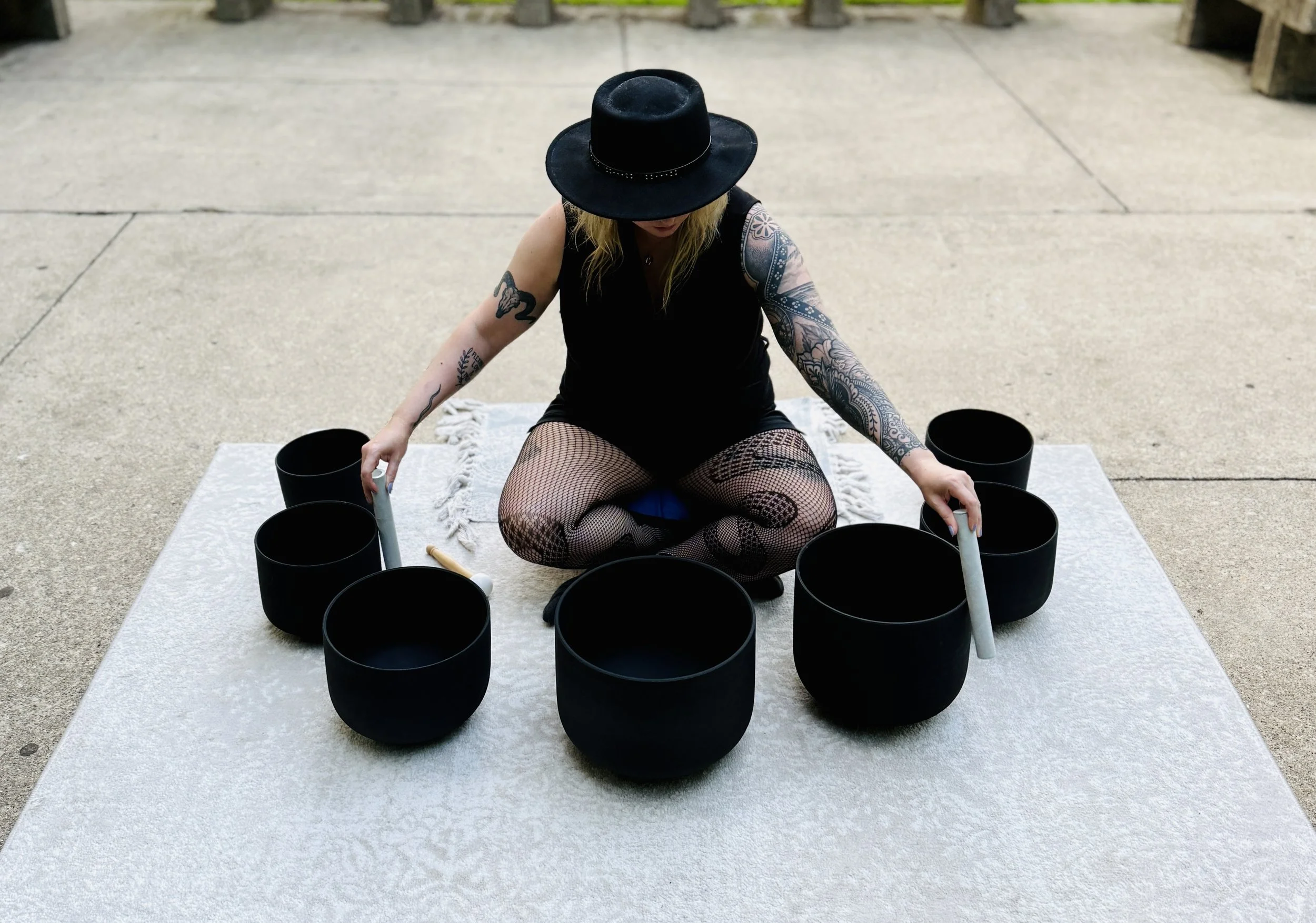 A woman sitting cross-legged on a gray rug on concrete pavement, playing black singing bowls with mallets. She has tattoos on both arms, wears fishnet stockings, a black sleeveless top, and a wide-brimmed black hat.