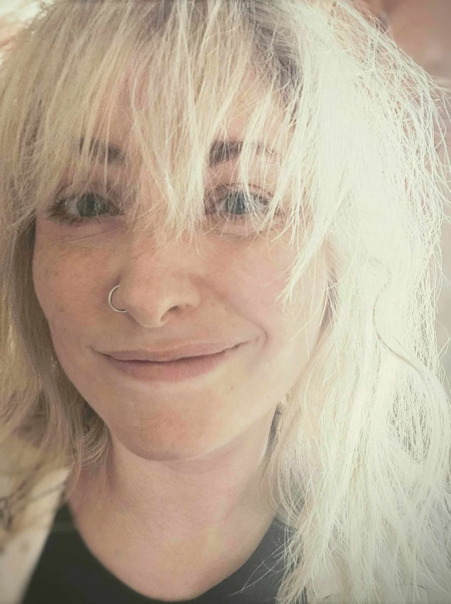 Close-up of a woman with long blond hair, wearing a nose ring, smiling softly.