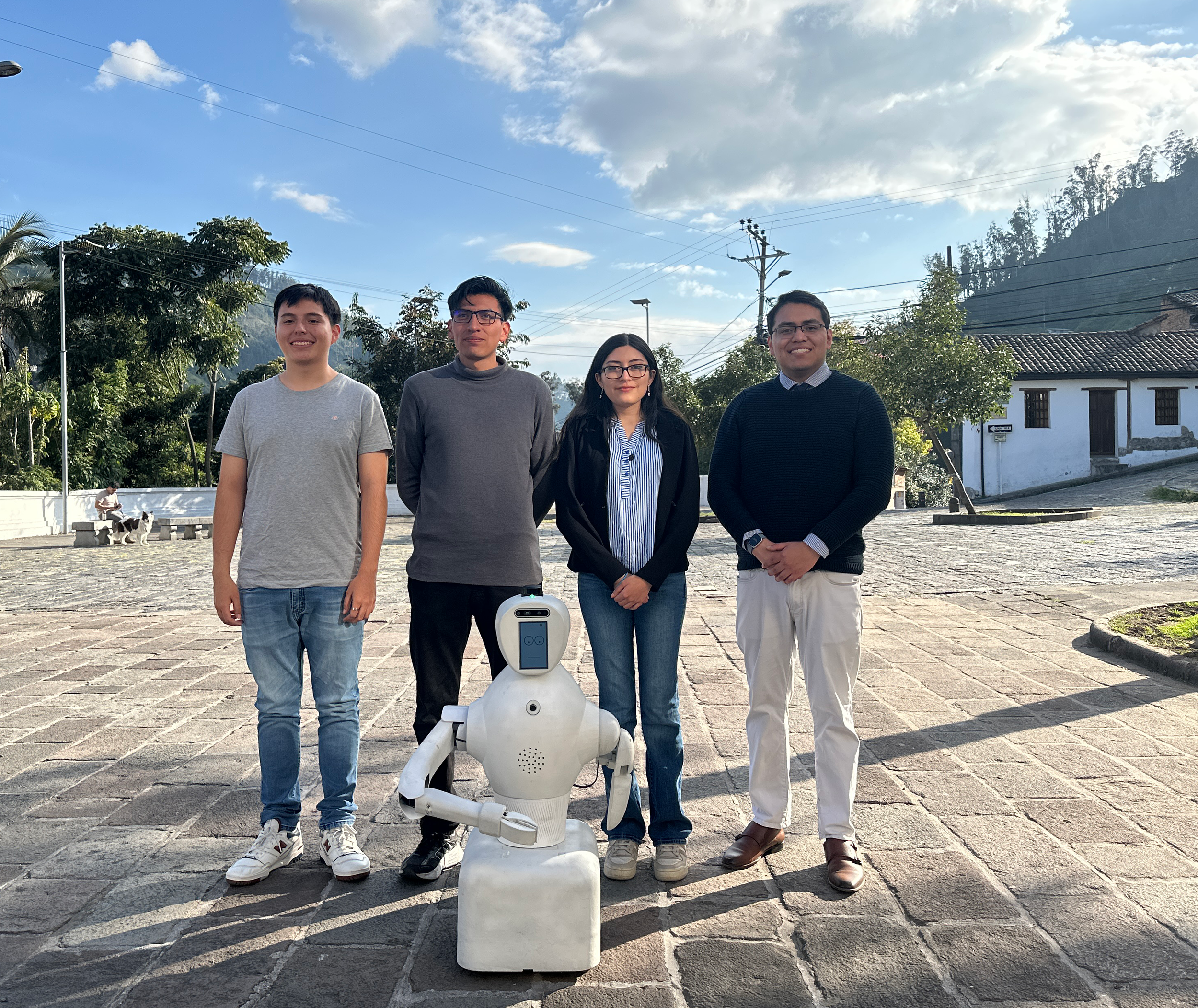 Four people standing outdoors in a row, with a white robot in front of them. The setting appears to be a town square with cobblestones, trees, and houses in the background, under a partly cloudy sky.