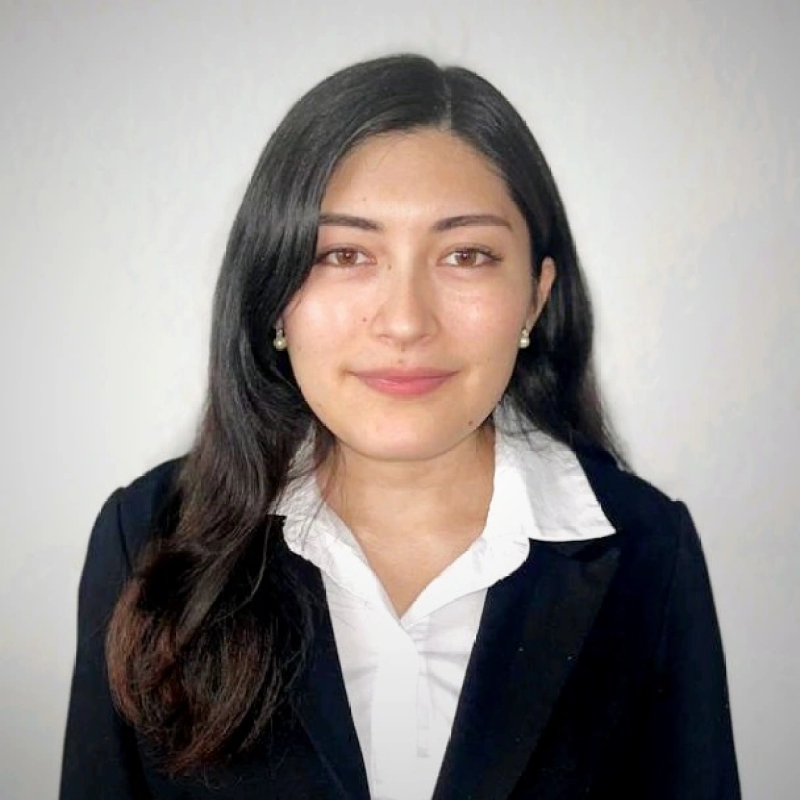 Portrait of a young woman with long dark hair, wearing a white shirt and black blazer, against a plain light-colored background.