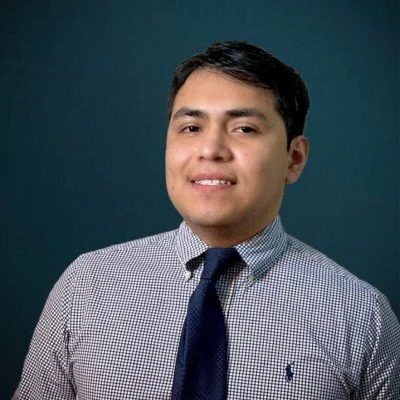 A young man in a checkered dress shirt and dark tie smiling against a solid dark background.