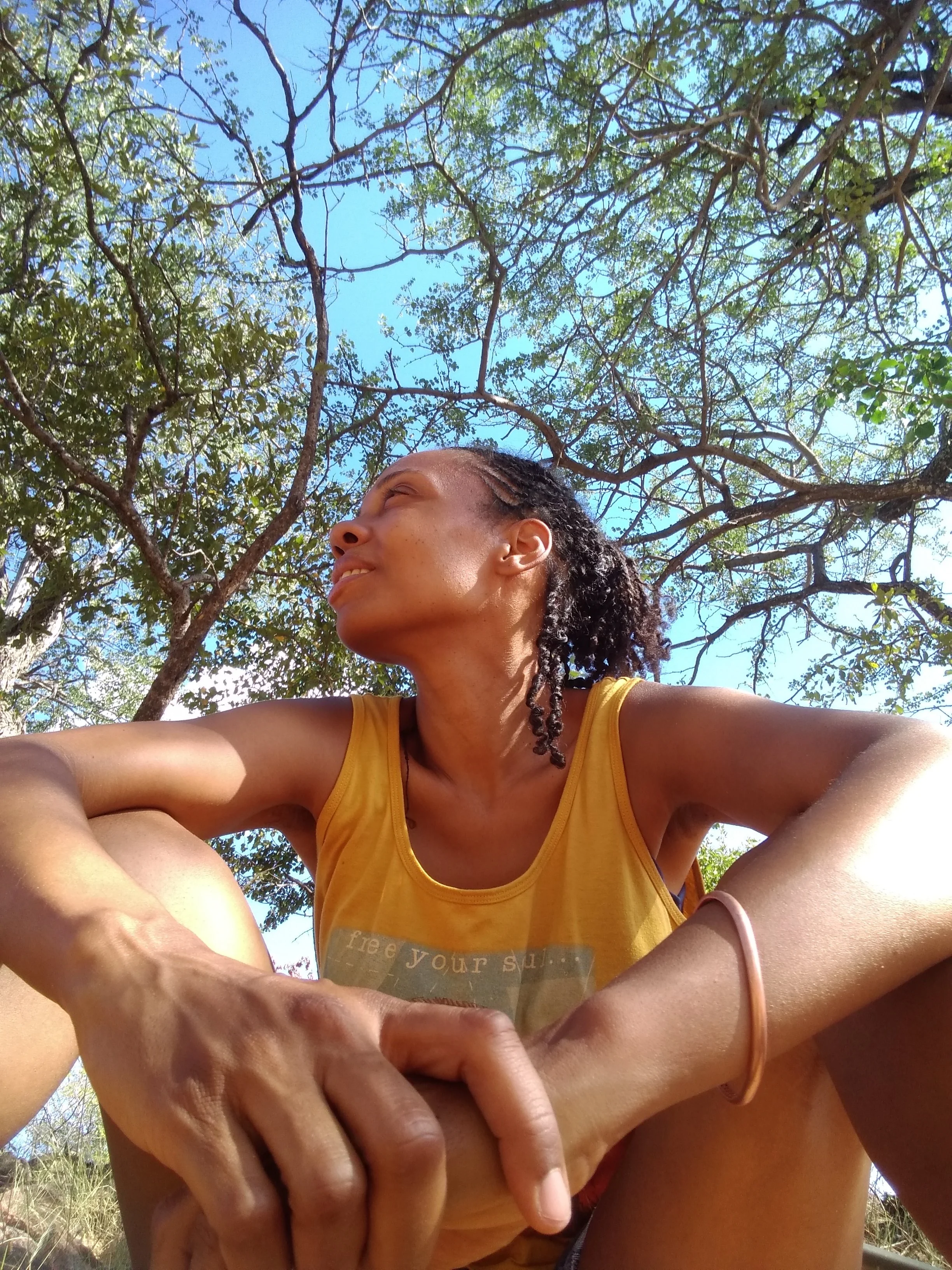 A woman sitting outdoors under a tree, wearing a yellow tank top, she feels content because she is using birth control without hormones.