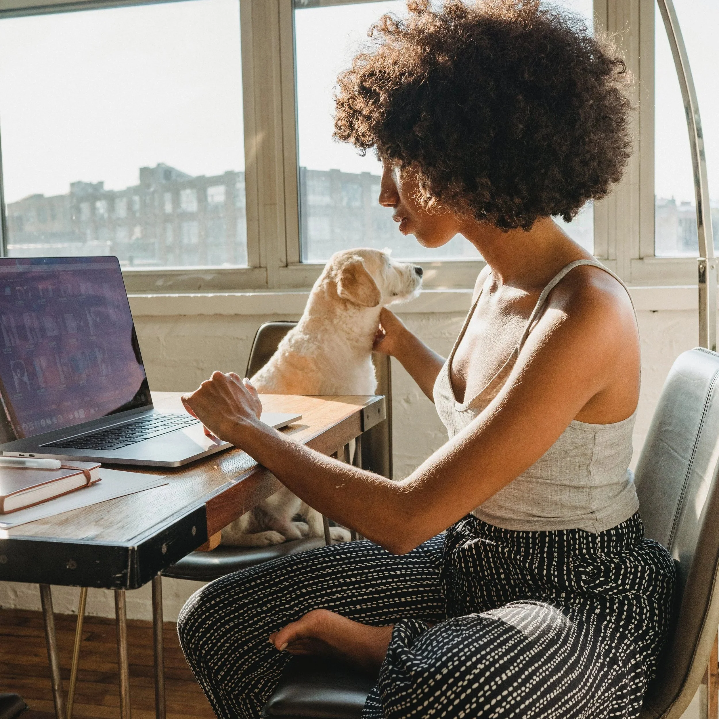 A woman with curly hair working on a laptop at a desk with a small white puppy sitting beside her, she is studying the fertility awareness method online.