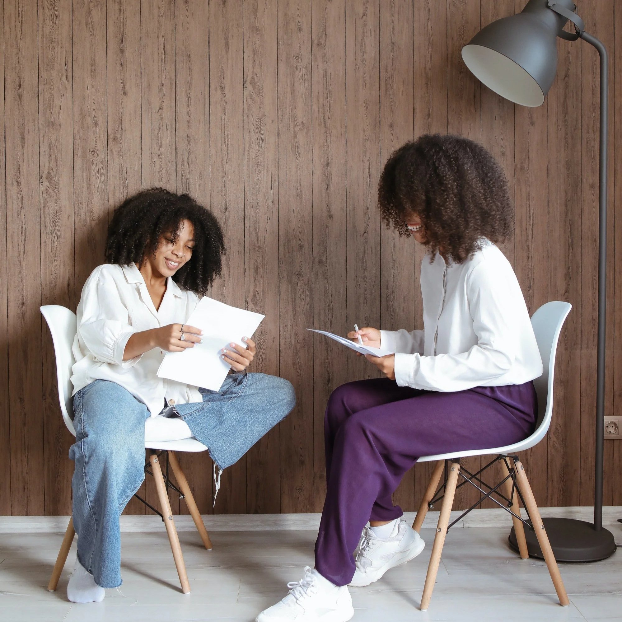 Two women with curly hair sit on chairs facing each other in a room with a wooden wall and a floor lamp, one is receiving personalized education on holistic women's health.