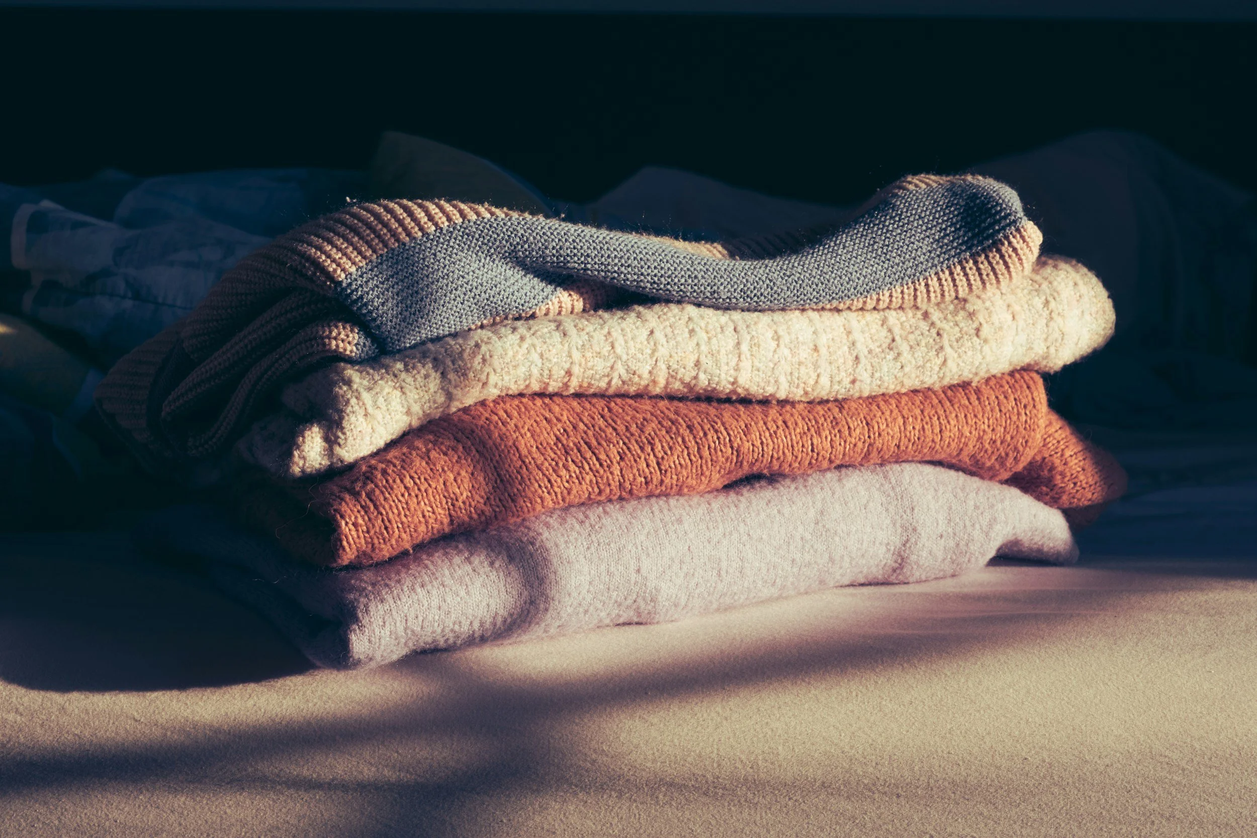 A neatly folded stack of cozy sweaters in muted tones, placed on a bed with a soft surface, with shadows cast across the foreground.