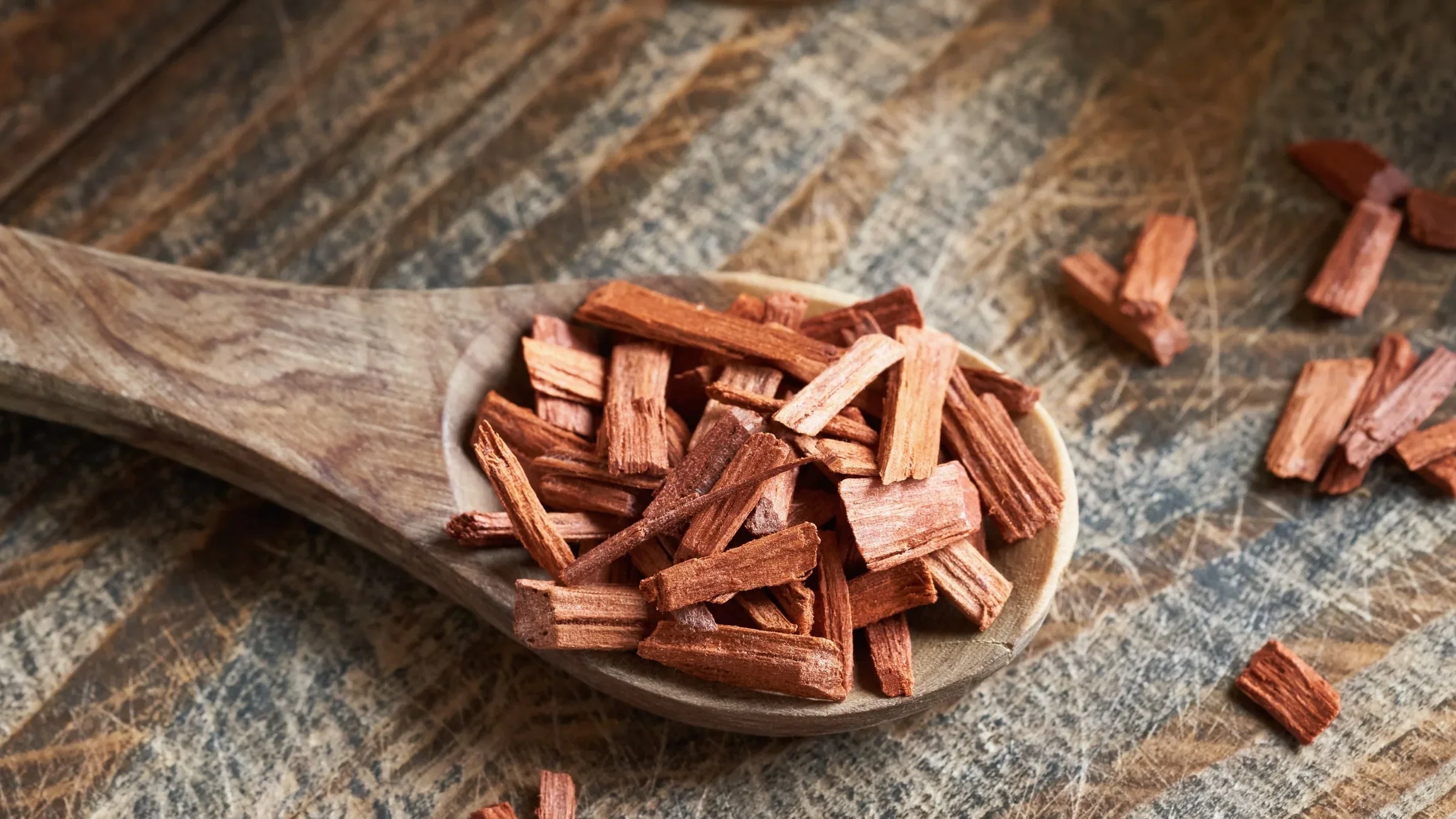 Close-up of a wooden spoon filled with dried pieces of sandalwood on a rustic wooden surface with a few sandalwood pieces scattered around.