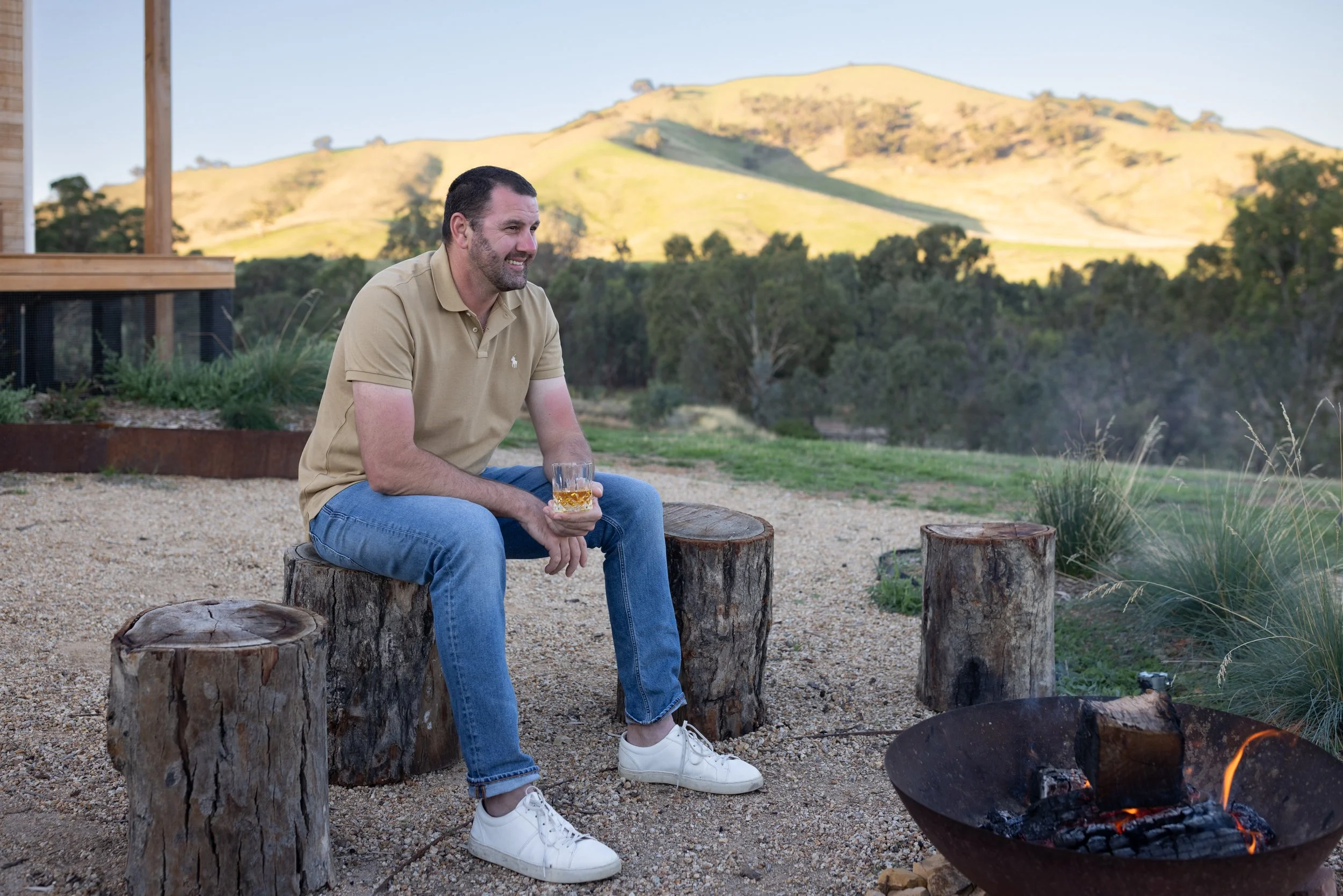 Man sitting on a log by a fire holding a glass of whisky in his hands with green mountains in the background.