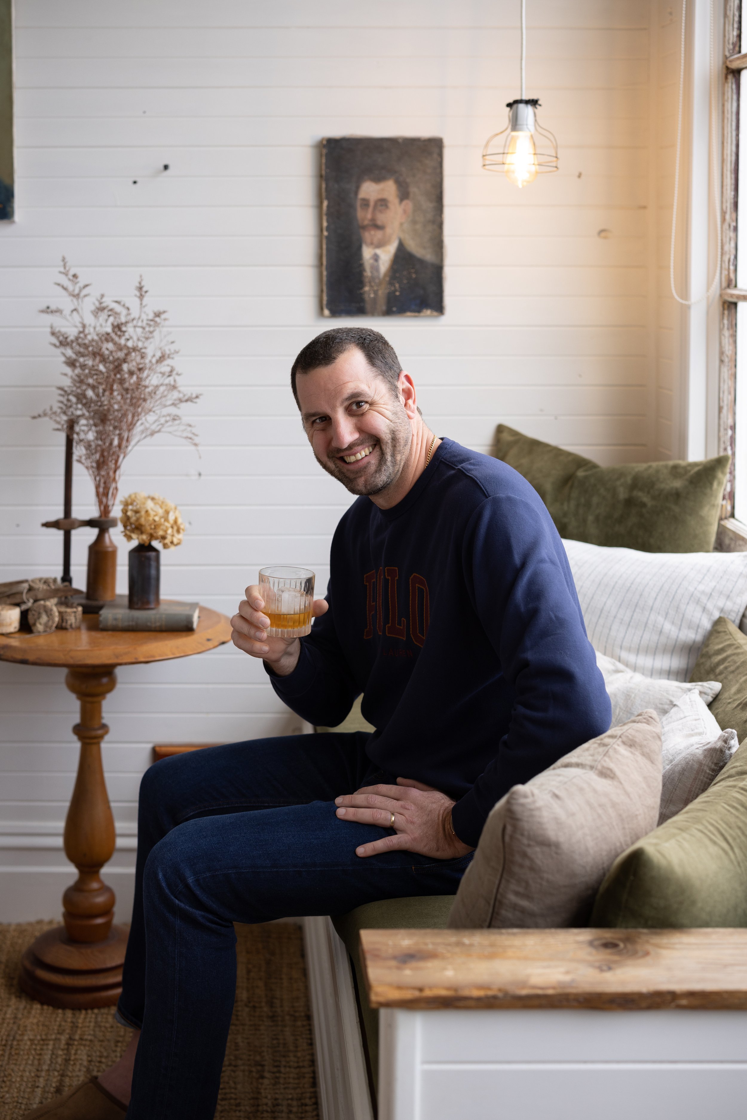 A smiling man with dark hair and a beard, wearing a navy blue sweatshirt, sitting on a sofa holding a glass of whiskey in his right hand, in a cozy living room with a wooden table, dried flowers, and art on the wall.