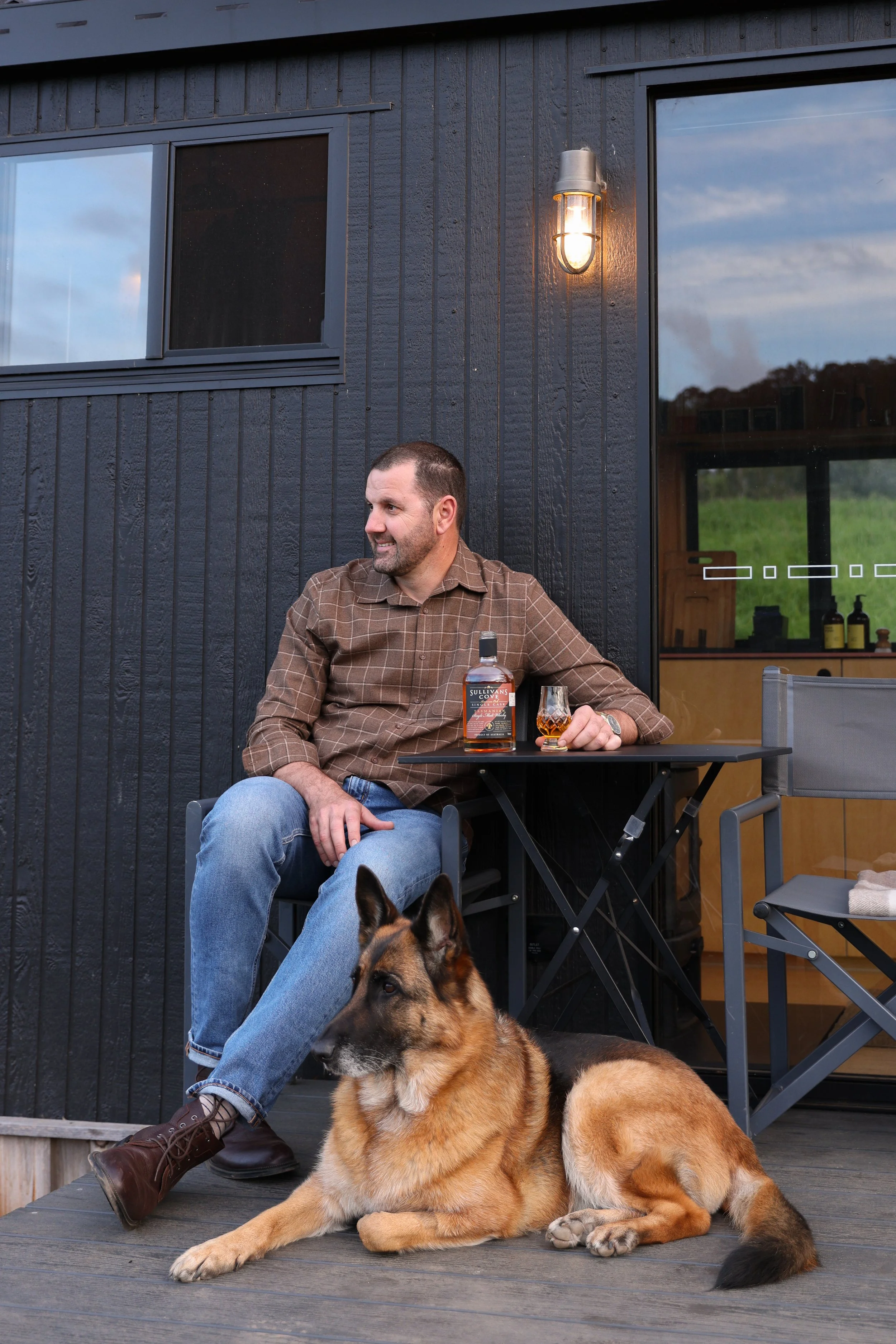 A man with a dog sitting on a wooden deck outside a black building, with a bottle of liquor and a glass of whisky on a small black table beside him.