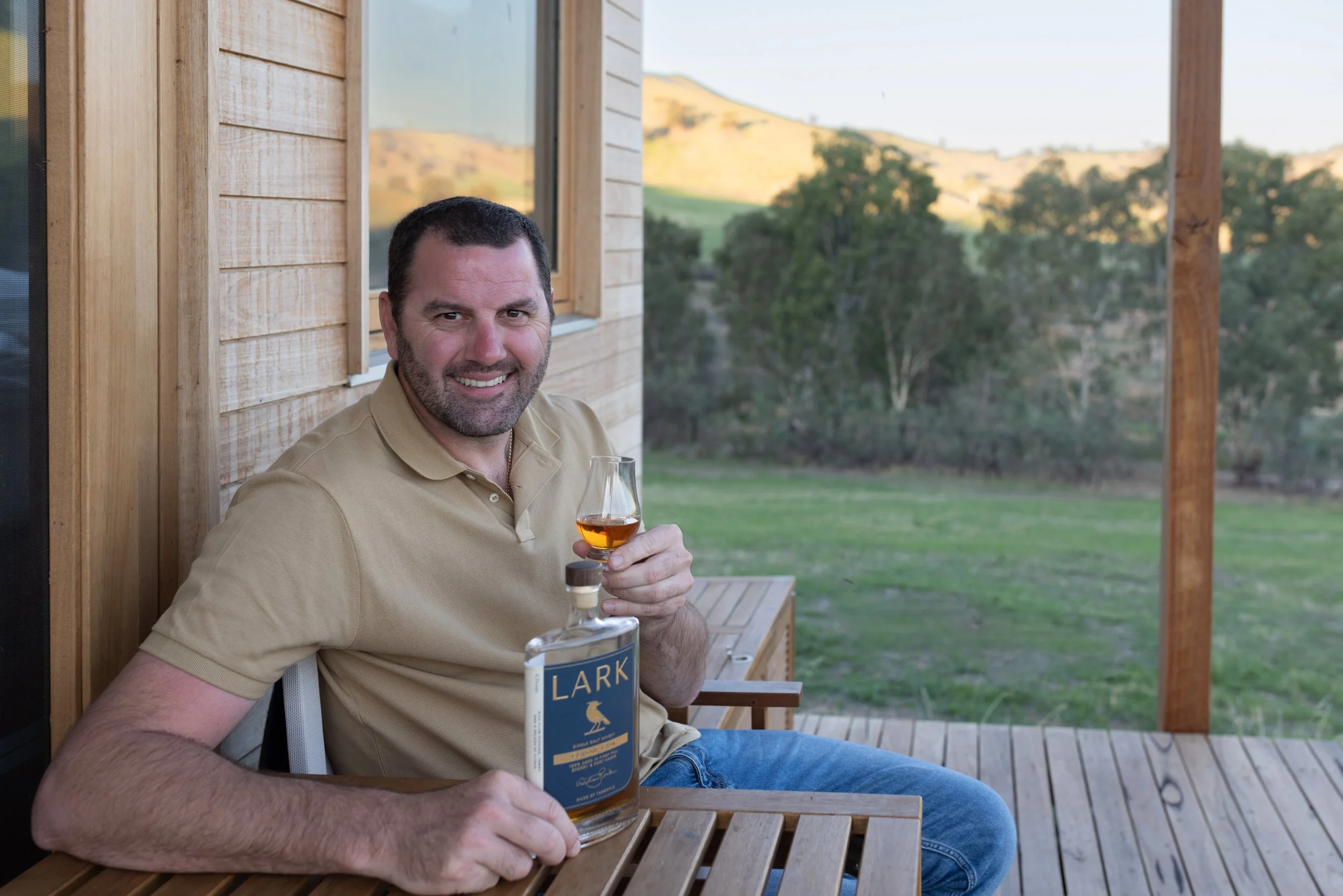 A man sitting on a deck smiling holding a glass of whisky and a bottle of Lark whisky.