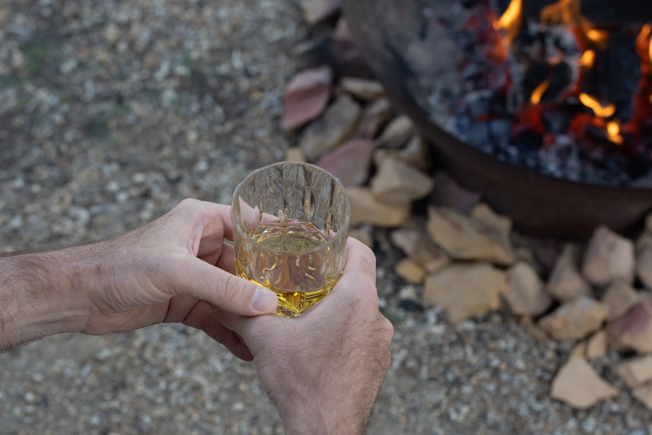Person holding a glass of whisky near a campfire with rocks around it.