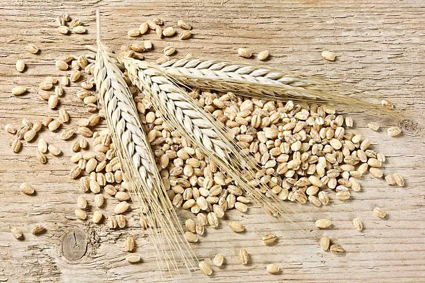 Three wheat stalks and scattered wheat grains on a wooden surface.
