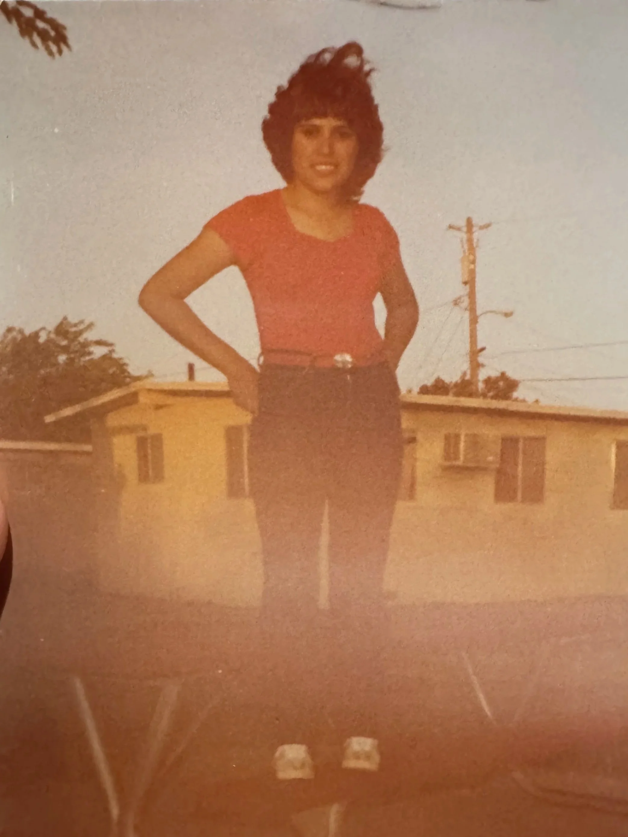 A woman with a large curly hairstyle wearing a red top, dark belt, and dark pants, standing outdoors on a balcony at dusk with an urban background including buildings, power lines, and a utility pole.