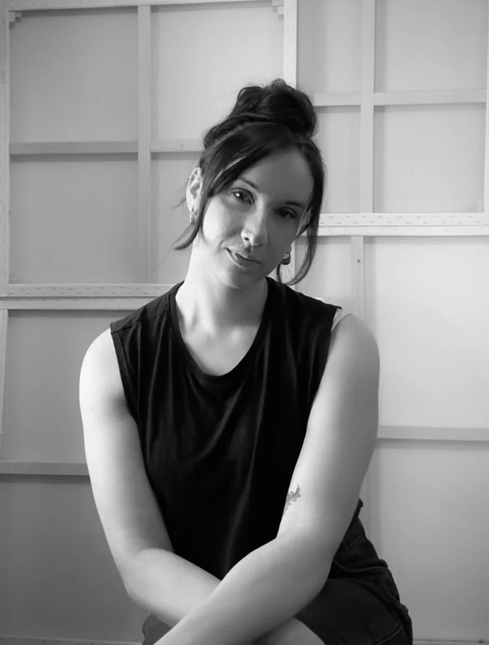 Australian female artist Savannah Mojidi sits in front of blank canvases, her hair is in a messy bun, she is wearing a black cropped tank top and dark black denim jeans, she is smiling