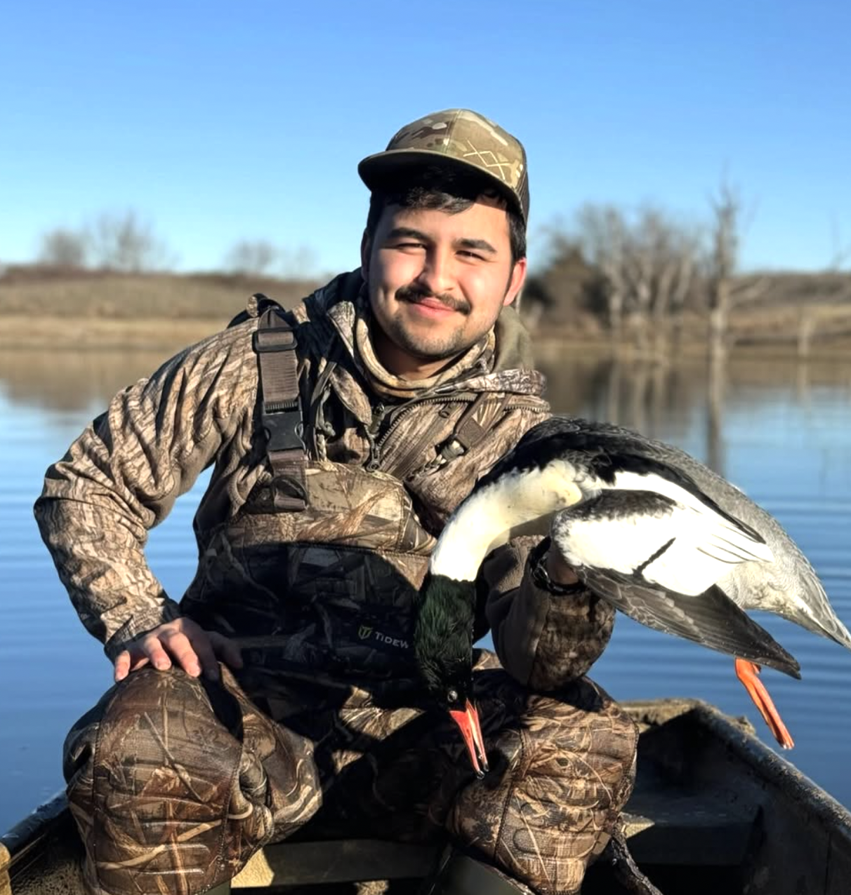 A man in camo outdoor gear holding a duck with a lake and distant trees in the background.