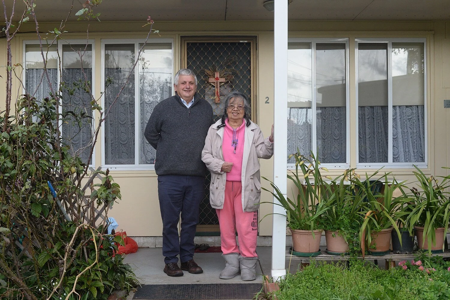 A man and woman stand in front of a home with pot plants and a garden.