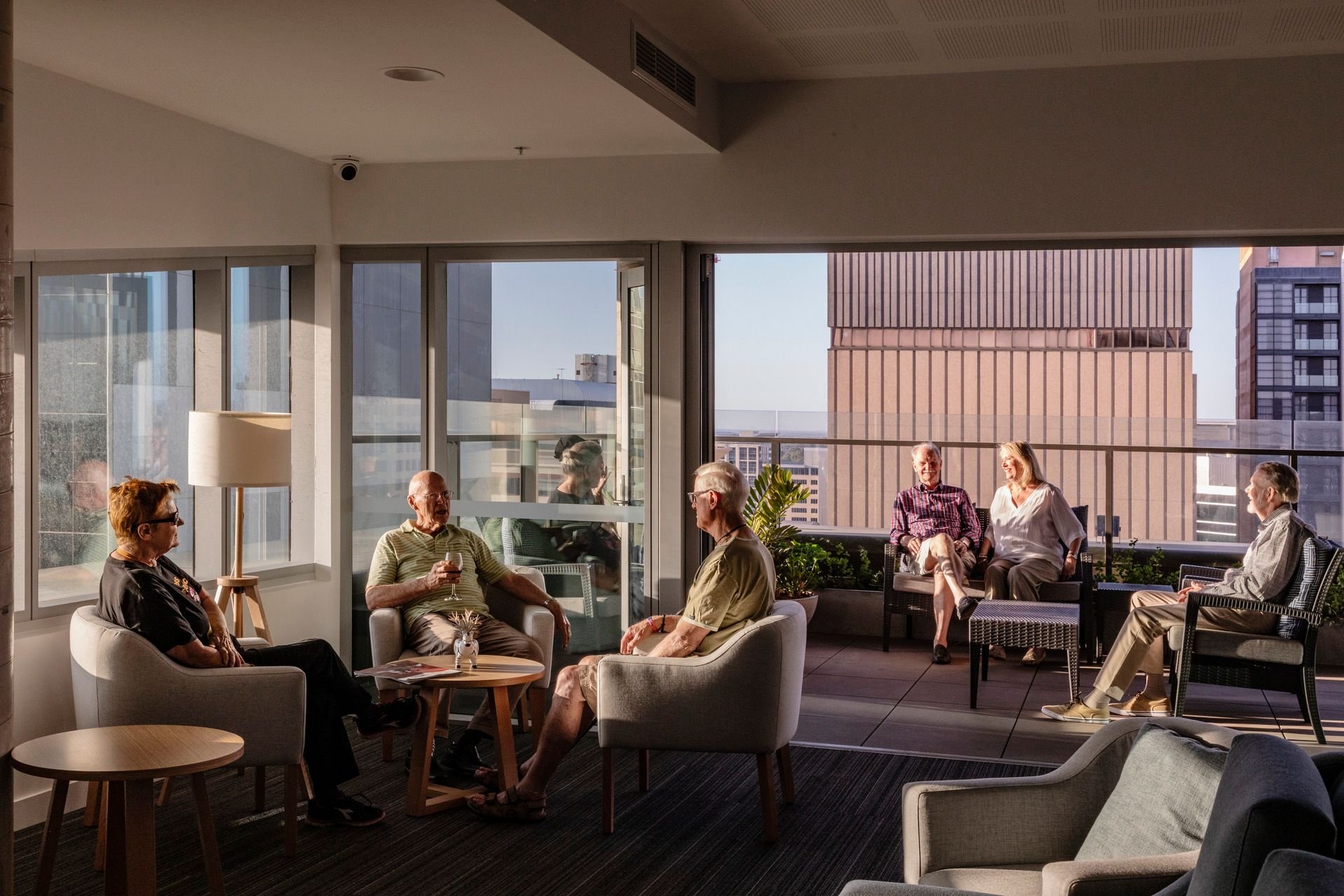 Communal living space showing interior spaces connecting to a large external balcony. People are seated together and talking.