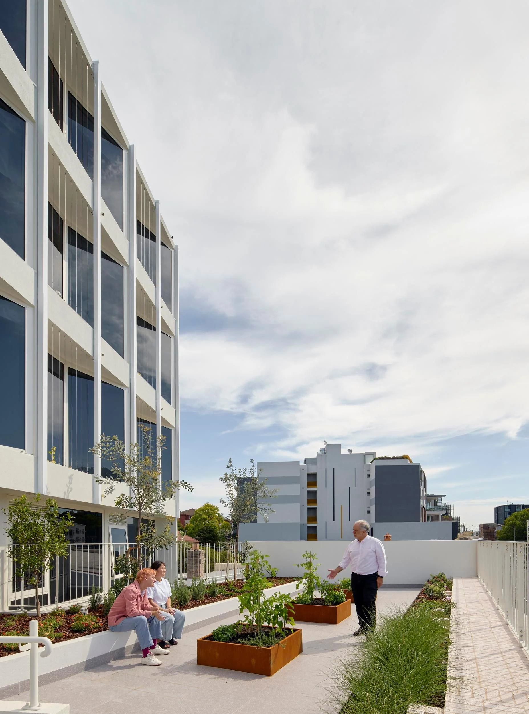 Communal outdoor terrace with people are seated and standing amongst planter boxes.