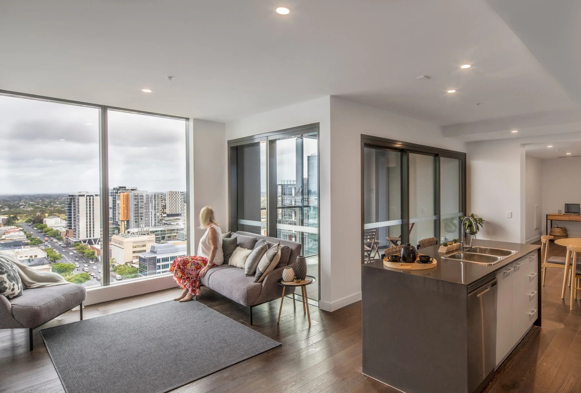 Retirement living apartment showing an open plan living, kitchen and dining area. A woman is sitting and looking at the view over the city.